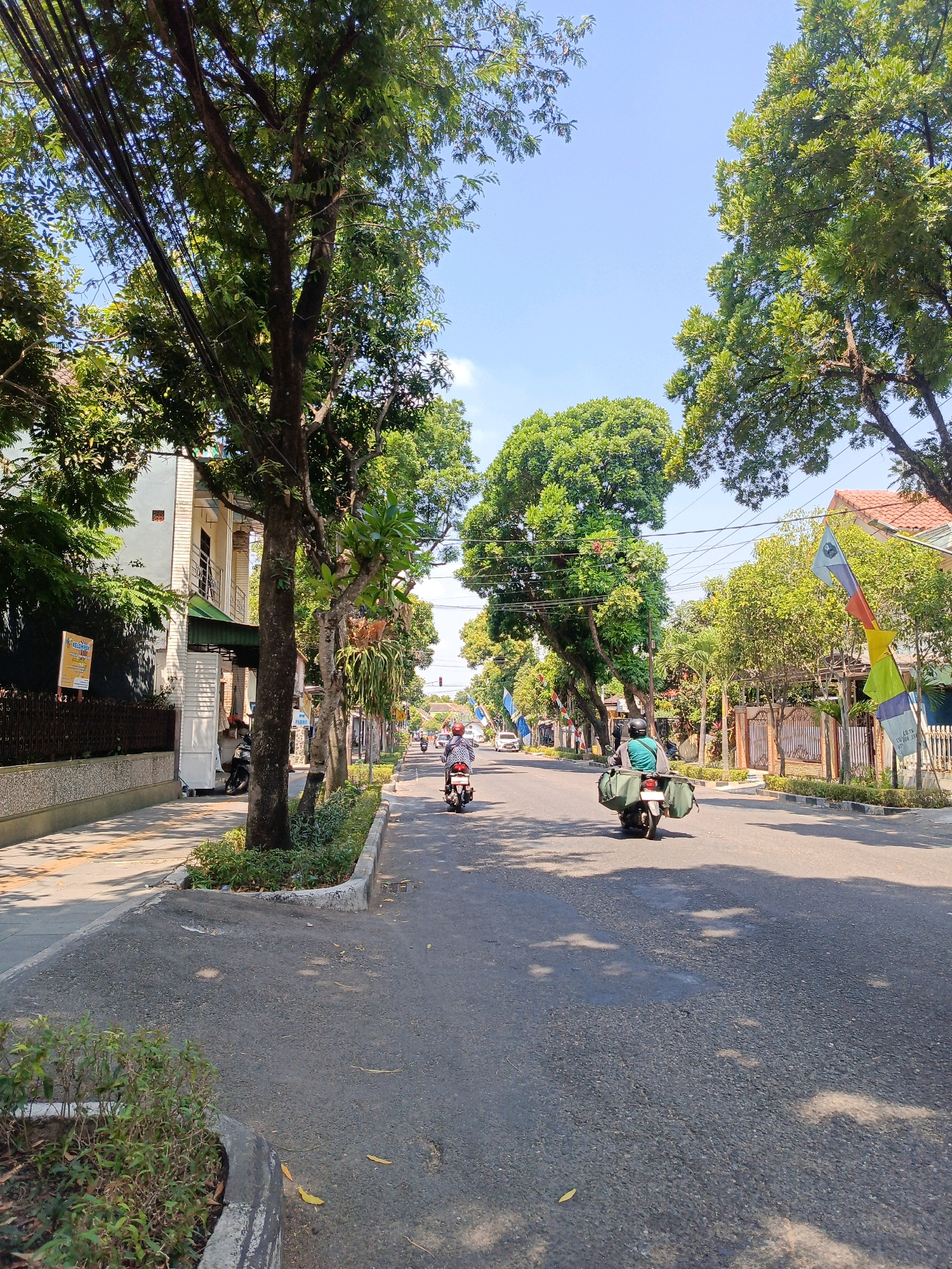 The image shows a street scene in Magetan, Indonesia, on a clear day. The street is paved and lined with trees on both sides. There are several motorcycles on the road, with riders wearing helmets. One motorcycle in the foreground has two large green bags attached to the back. On the left side of the street, there is a sidewalk with a building in the background. On the right side, there are houses with fences and flags. The overall impression is a typical residential street in a tropical climate.