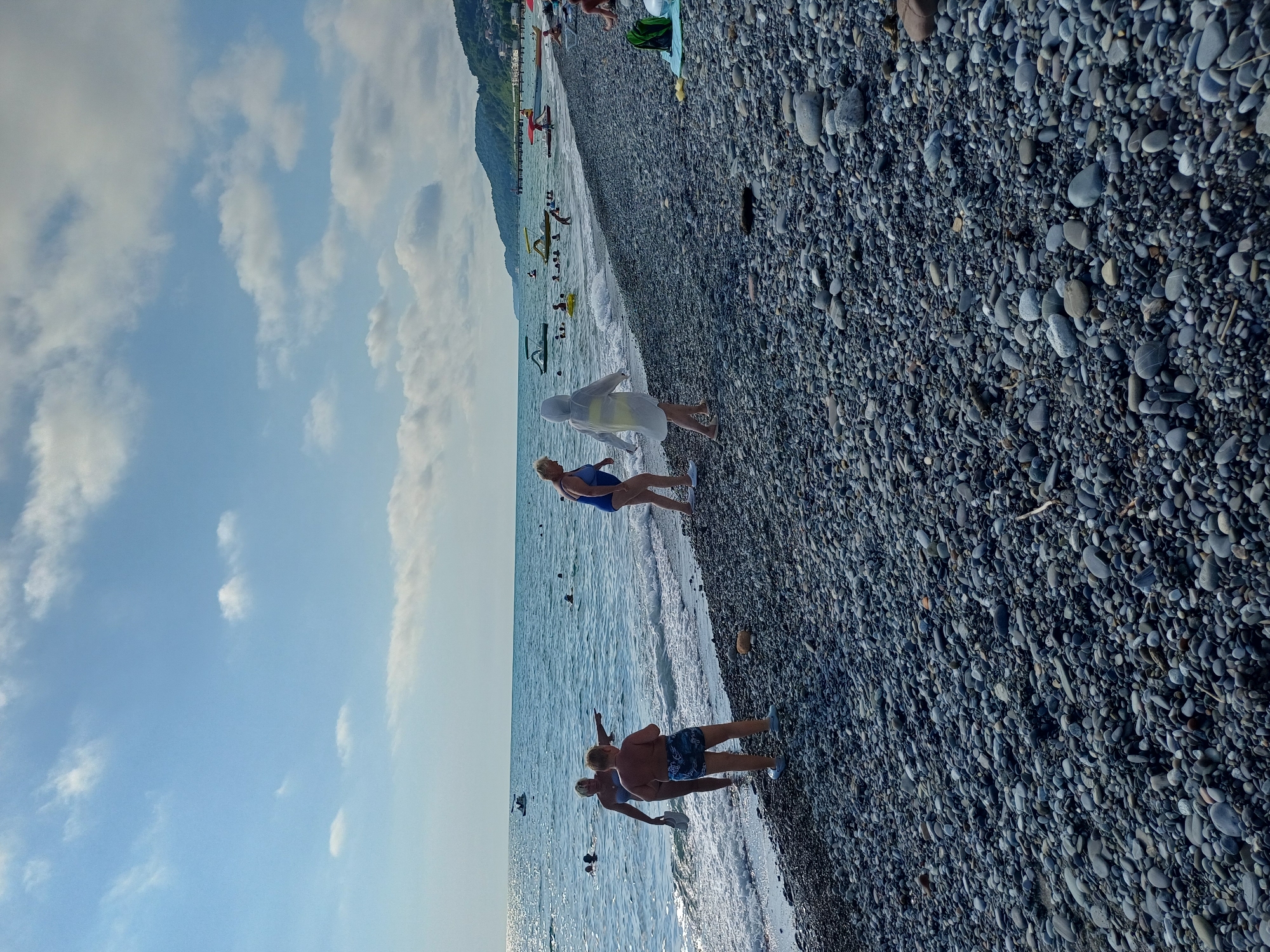 The image depicts a pebble beach on a sunny day in Sochi, Russia. The sea, with gentle waves, occupies the left side of the frame, reflecting the bright sky. A person in a blue swimsuit and another in a white hooded garment are visible on the shore. In the background, the sea is dotted with several boats, including what appear to be pedal boats, and a distant pier. The coastline is lined with lush green hills, and some buildings are visible on the slopes. The foreground is covered with an abundance of grey and black pebbles. The overall scene is one of a recreational beach environment.
