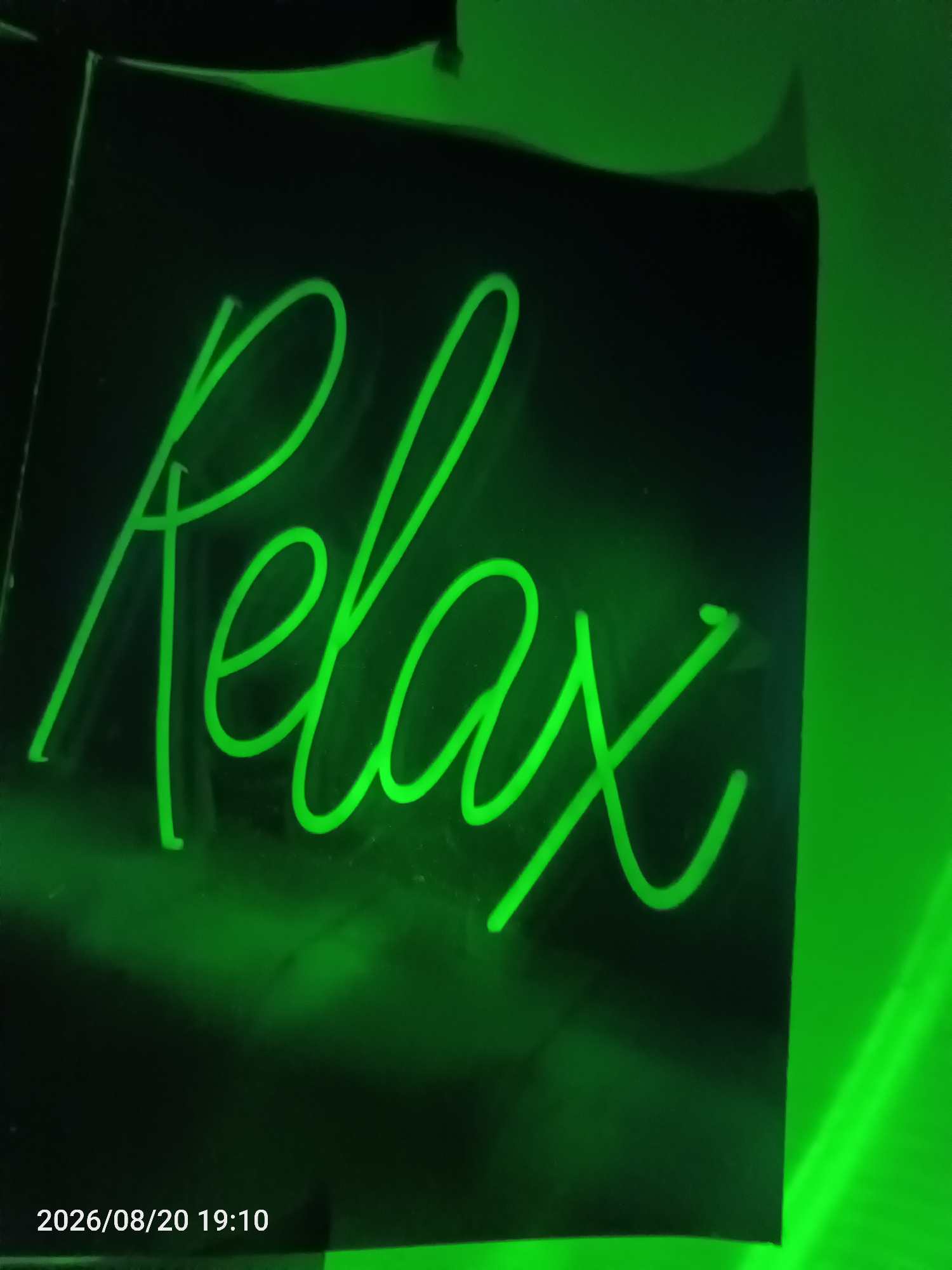 A neon sign shaped like the word "relax" glows green against a dark background. The sign is positioned indoors, possibly in a commercial establishment. The text "2026/08/20 19:10" is visible in the lower left corner, indicating the date and time the image was captured. The overall scene is illuminated by the vibrant green light of the neon sign, creating a somewhat ambient and relaxed atmosphere. No people are visible in the image. The location is identified as Jawilan, Indonesia.