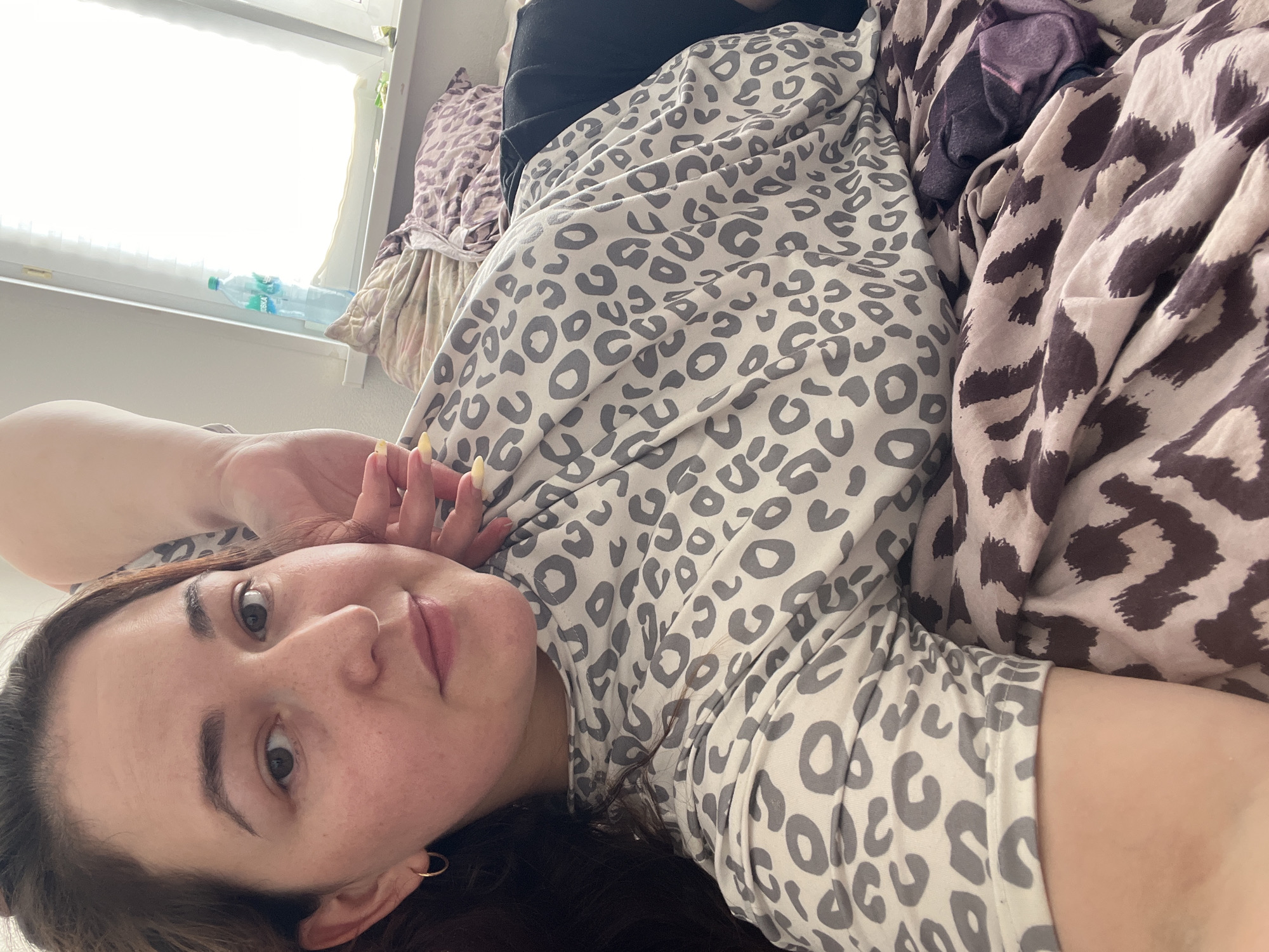 An overhead, close-up, selfie-style shot captures a young woman with dark brown hair, fair skin, and hazel eyes. She is lying down, propped up on her left arm, with her right hand resting on her cheek. Her mouth is slightly open, and she is looking at the camera. She wears a light grey t-shirt with a darker grey leopard print. Behind her, a window with white blinds lets in bright sunlight. On the windowsill, a plastic bottle of water is visible next to some pillows with a leopard print. The woman is positioned on a bed covered with a comforter featuring a brown and beige leopard print. The overall setting appears to be a bedroom in a residence within Chelyabinsk, Russia.