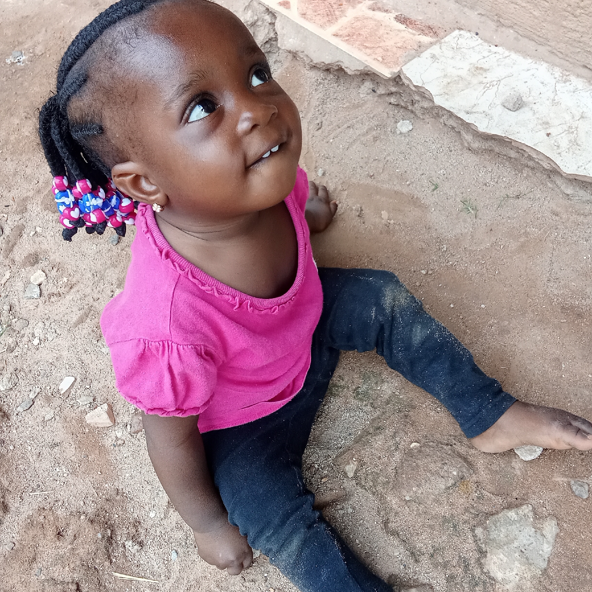 A young child sits on the ground, presumably in Akwanga, Nigeria. The child is Black, with braided hair adorned with colorful beads. She wears a pink shirt and dark leggings. The ground is sandy and uneven. The child looks upwards, her expression a mix of curiosity and delight. A small section of tile or brick is visible in the background.