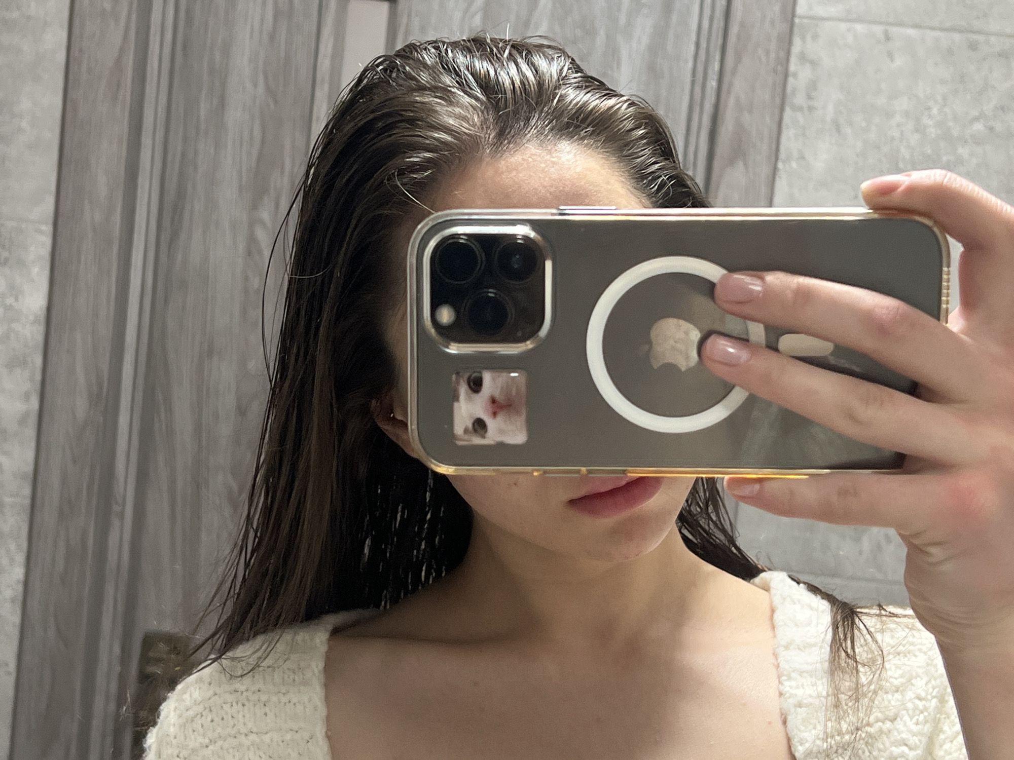 A person holds a smartphone in front of their face, partially obscuring it. The phone case is clear with a gold trim and a sticker of a cat near the camera lenses. The person has wet, dark brown hair and is wearing a white knit sweater. The background is a light gray wall, seemingly in a bathroom. There is no information to suggest the location is in Ternopil, Ukraine.