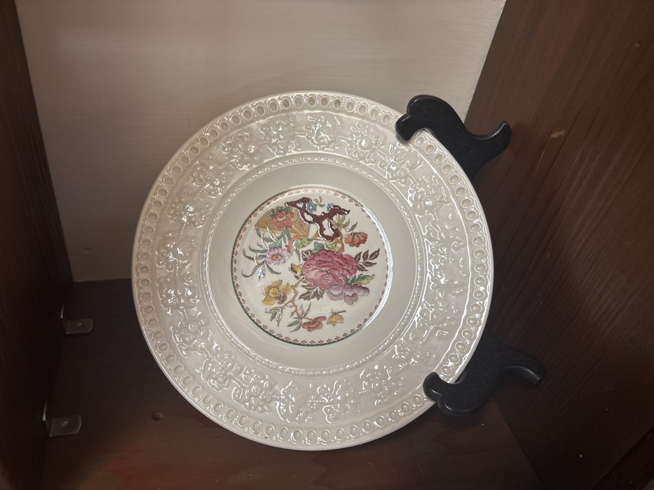 The image displays a cream-colored decorative plate, approximately 10-12 inches in diameter, positioned upright on a black, two-pronged plate stand. The plate features a central polychromatic floral design, including large pink blooms, smaller yellow and orange flowers, green leaves, and brown branch-like elements, enclosed by a thin green border. The wide rim of the plate is embossed with a repeating pattern of scrolled motifs and a continuous band of ovoid shapes. The plate and stand are situated on a dark brown wooden shelf or surface. The immediate background consists of dark brown wooden walls on the left and top, with a lighter-toned wall on the right. Two silver-colored L-brackets are visible in the upper left corner of the wooden enclosure. No individuals are present, and no active interactions are depicted. The specific location within the city of Monterey, United States of America, cannot be determined from the visual evidence provided.
