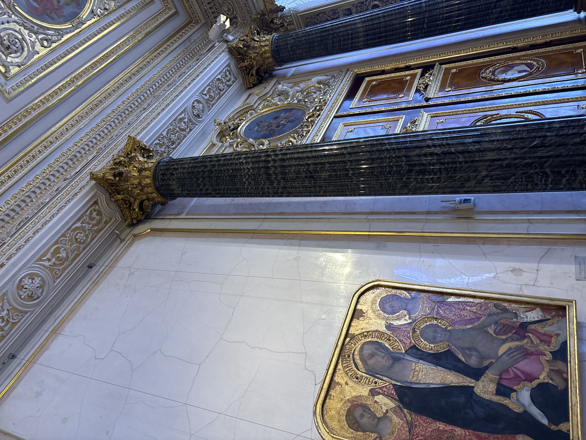 The image depicts an interior view of the Church of the Savior on Spilled Blood in Saint Petersburg, Russia. The scene is characterized by ornate architecture, featuring gilded decorative elements, marble columns, and intricate ceiling moldings. On the left wall, a framed religious painting of the Virgin Mary and child is prominently displayed against a cracked, light-colored marble surface. To the right, a large, dark marble column with a gilded capital stands tall, partially obscuring a set of wooden doors adorned with golden accents and oval portraits. The overall impression is one of opulent religious grandeur.