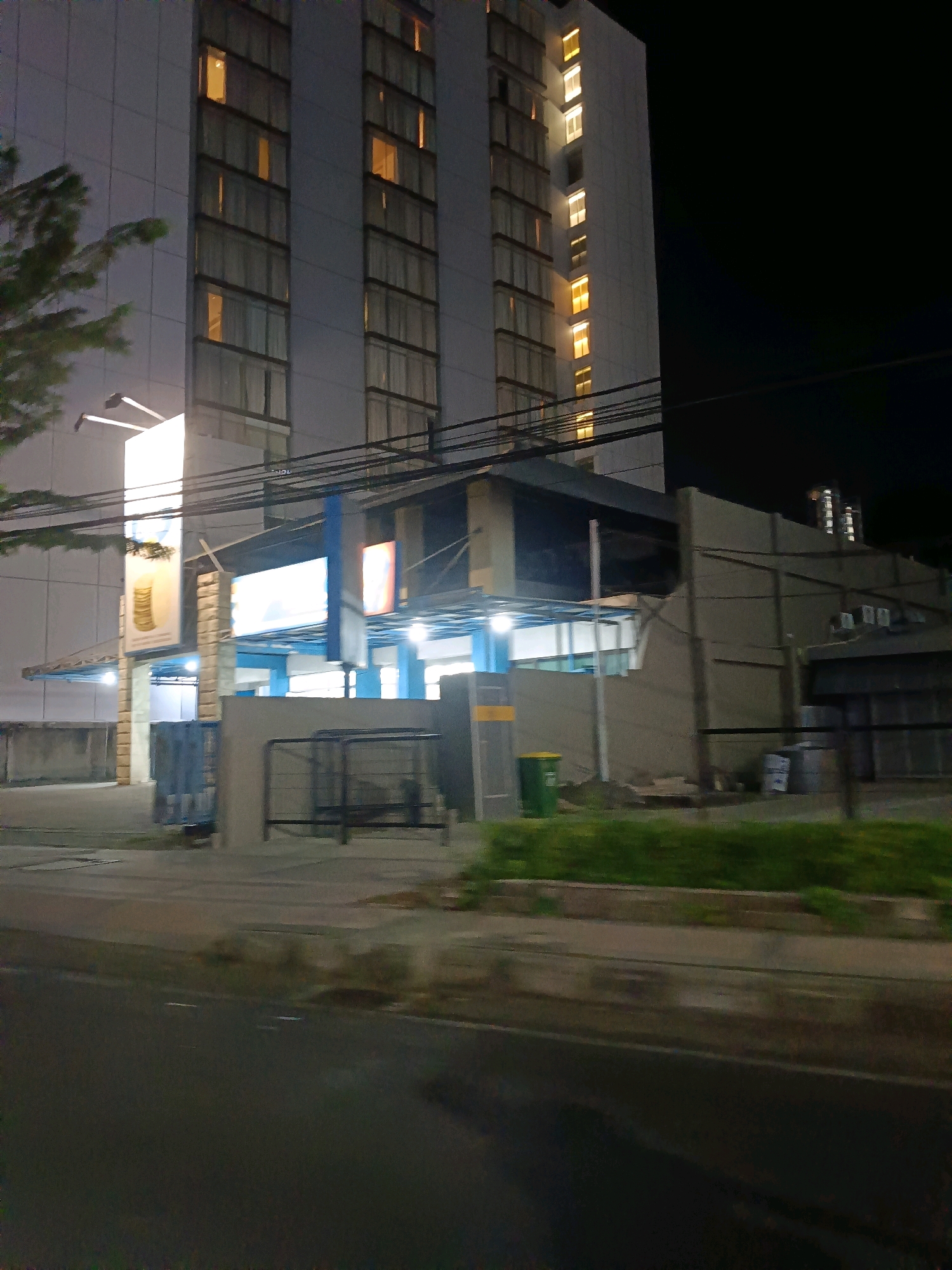 The image captures a nighttime view of a tall, multi-story building, likely a hotel or office building, located in Surabaya, Indonesia. The building's facade is predominantly white, with numerous windows illuminated from within by a warm, yellow light, suggesting that some floors or rooms are occupied. A portion of a tree with dark green foliage is visible on the left side of the frame, silhouetted against the brightly lit building.
At the base of the building, a lower structure with a dark roof and blue accents is visible. This area appears to be an entrance or a lobby, with illuminated signs displayed. One prominent sign on the left features an image of stacked gold coins. Another sign, to the right, is a large, brightly lit rectangular display. A concrete wall with a dark metal fence or gate separates the building's immediate area from the foreground. A green trash bin is positioned near this wall.
The foreground consists of a paved area, likely a sidewalk or parking lot, leading to a street or road, which is wet, indicating recent rain or damp conditions. The street is dark and shows faint white markings. Power lines and cables are visible crisscrossing the scene above the lower structure. In the background, the night sky is dark, with a few distant lights suggesting other urban structures. The overall scene depicts a typical urban environment at night, with the building serving as the central point of focus.
