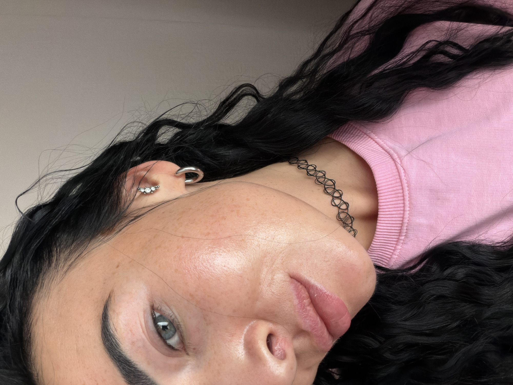 A close-up portrait shows a woman with fair skin, freckles, and black curly hair wearing a pink top and a black choker. She has light blue eyes and is wearing earrings. The background is a plain, light-colored wall. The image is angled. This image does not appear to be in Saint Petersburg, Russia.