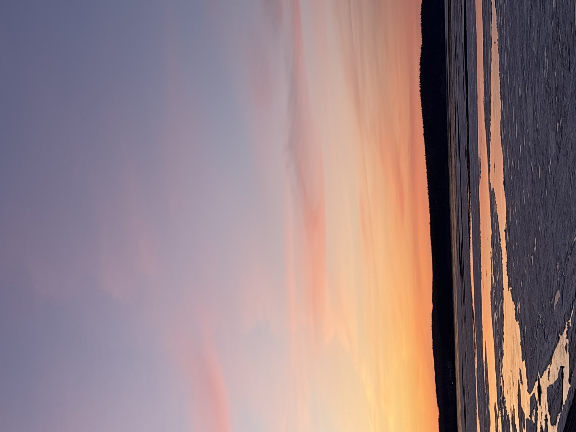 This landscape image captures a serene sunrise or sunset over a wide body of water in Chaikovsky, Russia. The sky dominates the upper portion, displaying a striking gradient of colors. On the left, it transitions from a soft, cool lavender-blue into warmer pinks and oranges towards the center and right, where the sun's direct light paints the clouds in vibrant golden and amber hues. Wispy, elongated clouds are visible, adding texture to the colorful expanse.
A dark, silhouetted landmass, likely a distant treeline or shoreline, stretches across the horizon, separating the sky from the water. The water below reflects the brilliant colors of the sky, creating shimmering streaks of orange, yellow, and pink across its surface. The surface of the water shows textural details, appearing somewhat uneven or partially covered, possibly suggesting ice or a disturbed surface. The low angle of the sun, indicated by the intense colors and long reflections, confirms it is either dawn or dusk. The overall impression is one of natural beauty and tranquility. There is no visible text in the image.