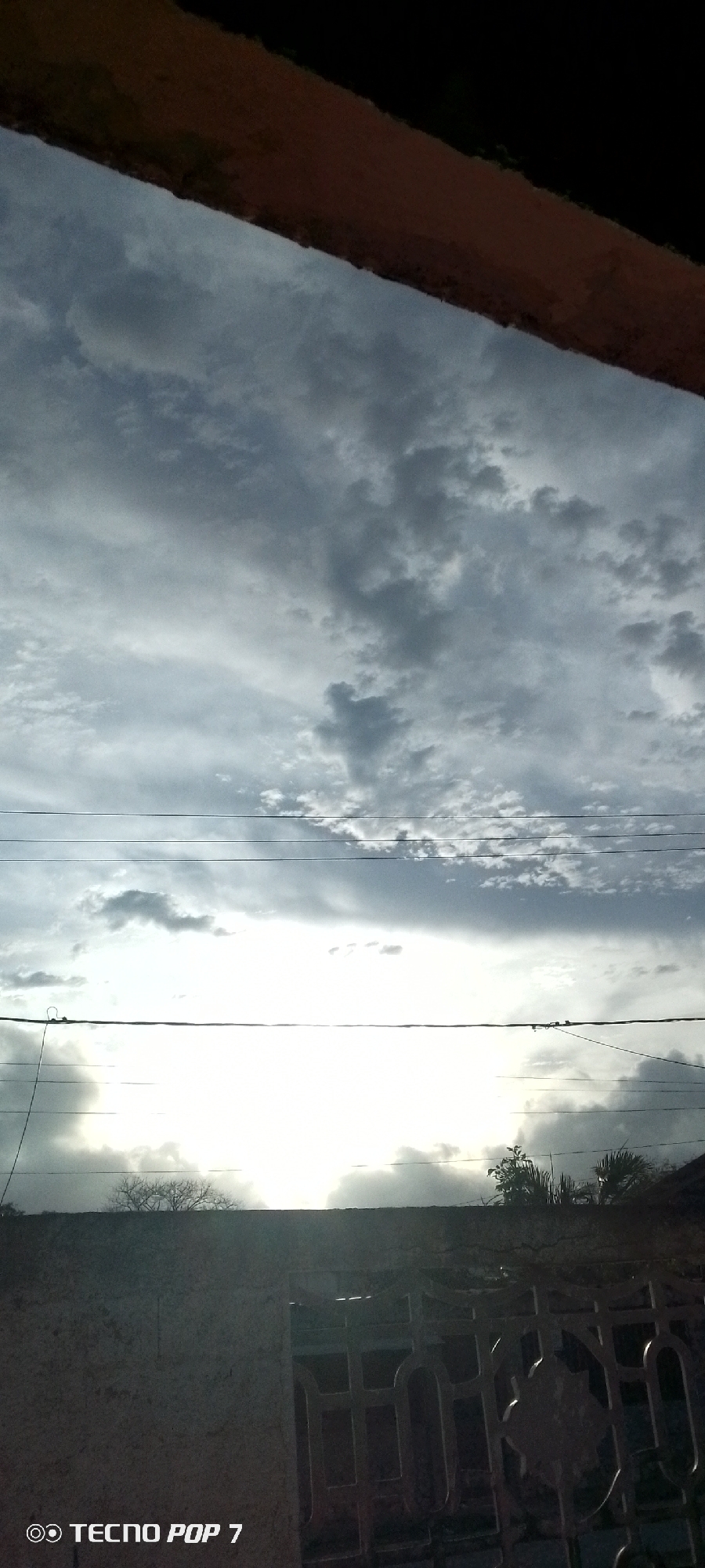 The image shows a cloudy sky with visible power lines. Below the sky, there's a partial view of a wall and a metal gate. There are trees and other vegetation behind the wall. The top of the image includes part of a building structure, likely a roof or a building edge. The text "TECNO POP 7" is visible in the lower left corner. The photo appears to have been taken in Altagracia, Venezuela.
