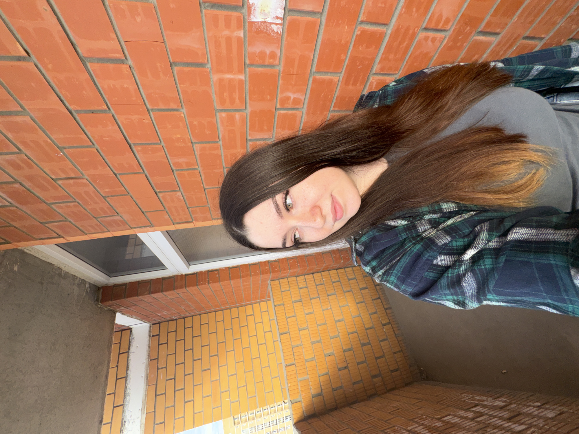 A young woman with long brown hair is taking a selfie outdoors. She is wearing a grey t-shirt and a green and blue plaid shirt over it. She has light skin with some freckles and is looking directly at the camera with a slight smile. Her eyes are green and she has dark eyebrows. The background consists of brick walls. To her left, there is a red brick wall, and below that, a window with a white frame. To her right and below, there is a yellow brick wall. In the distance, several apartment buildings are visible. The lighting suggests it might be daytime. The scene appears to be an outdoor architectural space, possibly a balcony or courtyard under construction. The overall mood is casual and relaxed. No visible text is present in the image.