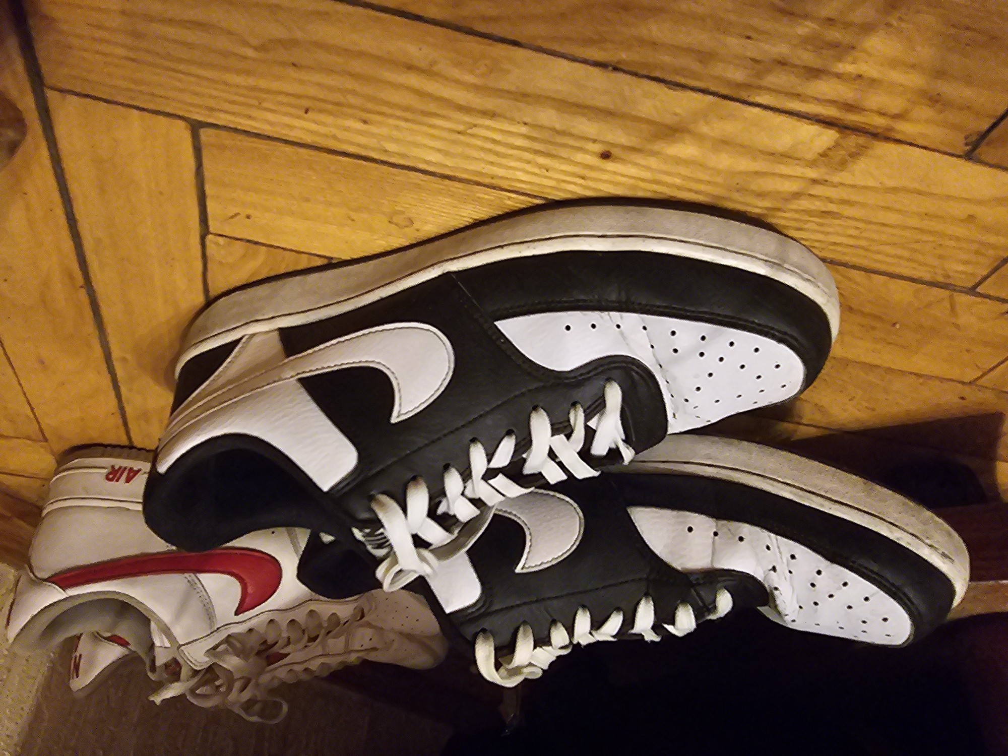 This image shows a pair of black and white Nike sneakers in the foreground, positioned on a wooden floor. Behind them, another pair of white Nike sneakers with red accents is visible. The sneakers appear to be worn. The environment is indoors, likely a room with natural lighting, as suggested by the shadows on the wooden floor. There are no people in the image. The scene is a close-up shot of footwear, with no discernible landmarks or features that would indicate a specific location in Vilnius, Lithuania.