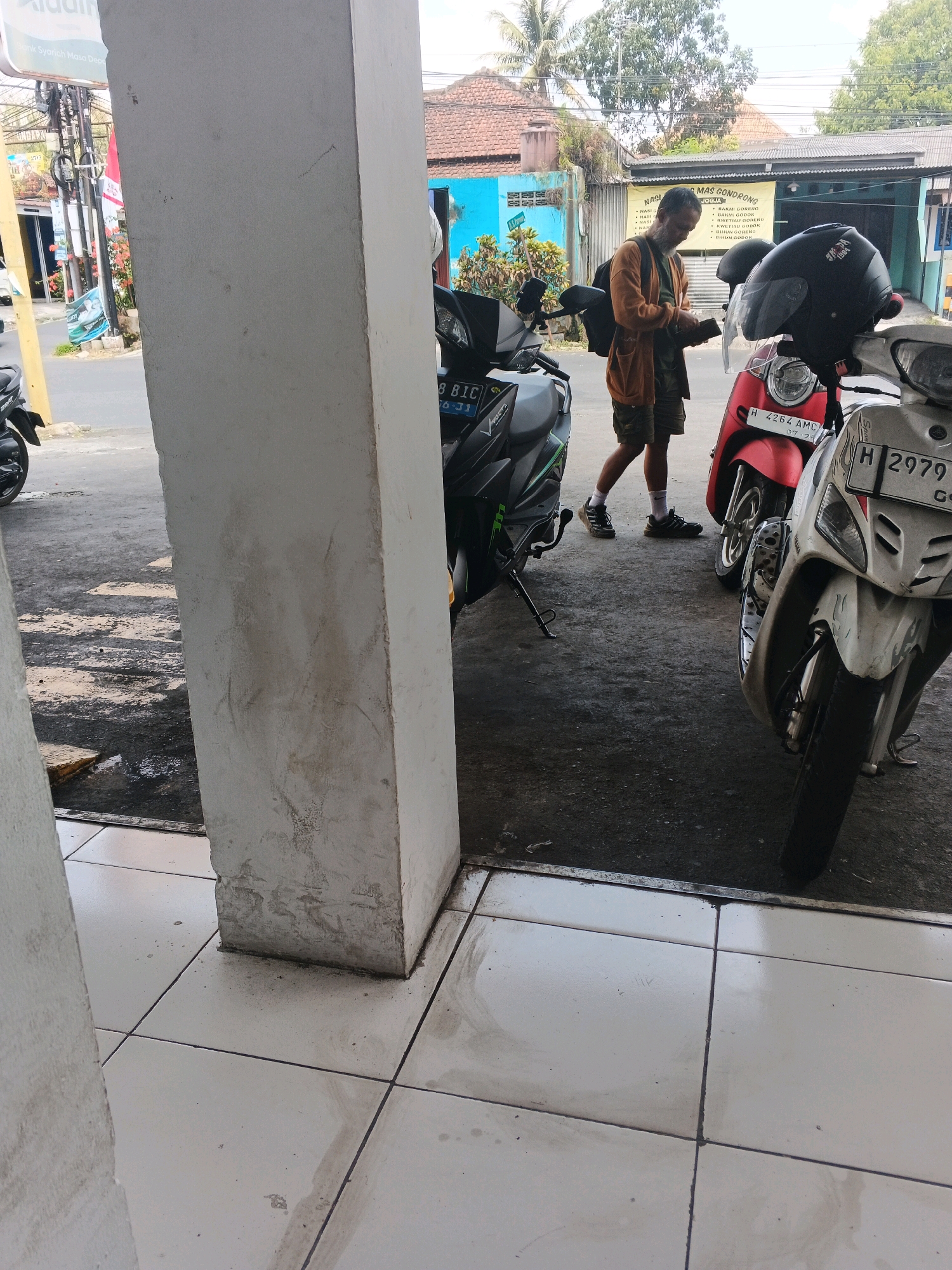 A man stands near several motorcycles in Sidomulyo, Indonesia, checking his wallet. The man is wearing a brown cardigan, a green t-shirt, and camouflage shorts. He is standing next to a red scooter and a white scooter, with a gray motorcycle to his left. The man is looking down at his wallet and appears to be engaged in a transaction or decision-making process. The scene is likely a street or parking area in front of a building with a sign that reads "NASI MAS GONDONG JOGJA." The ground is paved and somewhat wet, with tiles in the foreground.