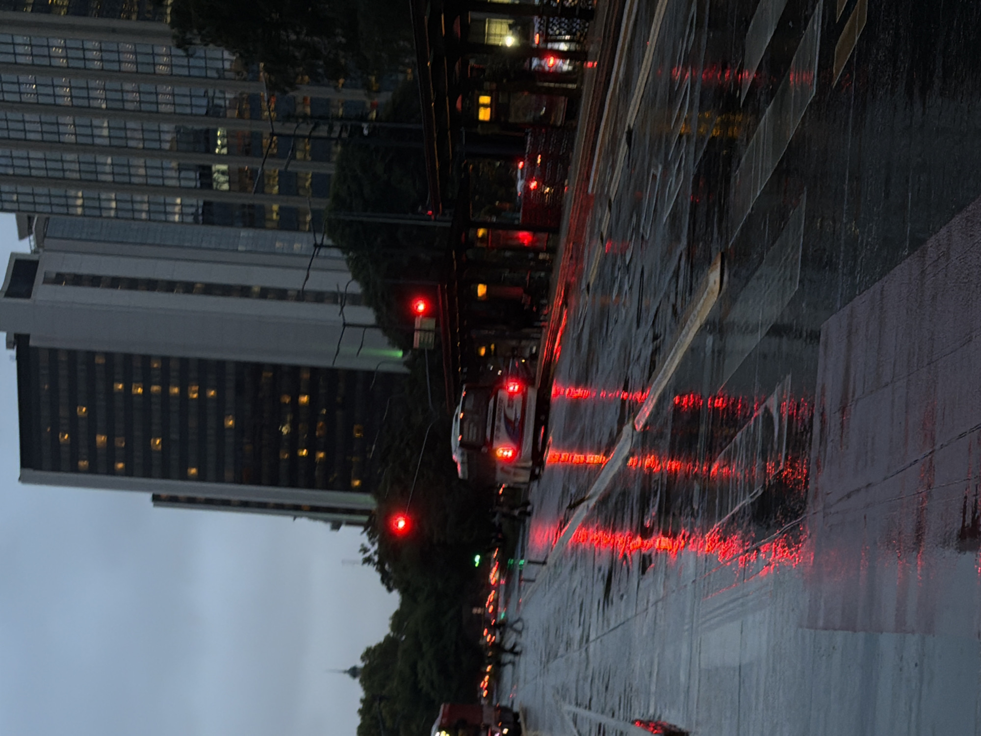 This is an image taken at nighttime on a rainy street in Buenos Aires, Argentina. Tall buildings dominate the left side of the frame, suggesting a downtown area. Reflections of red taillights and streetlights streak along the wet pavement, indicating the presence of traffic. A bus is visible amidst the lights. Pedestrians are silhouetted, walking along the sidewalk. The sky is overcast and gray.