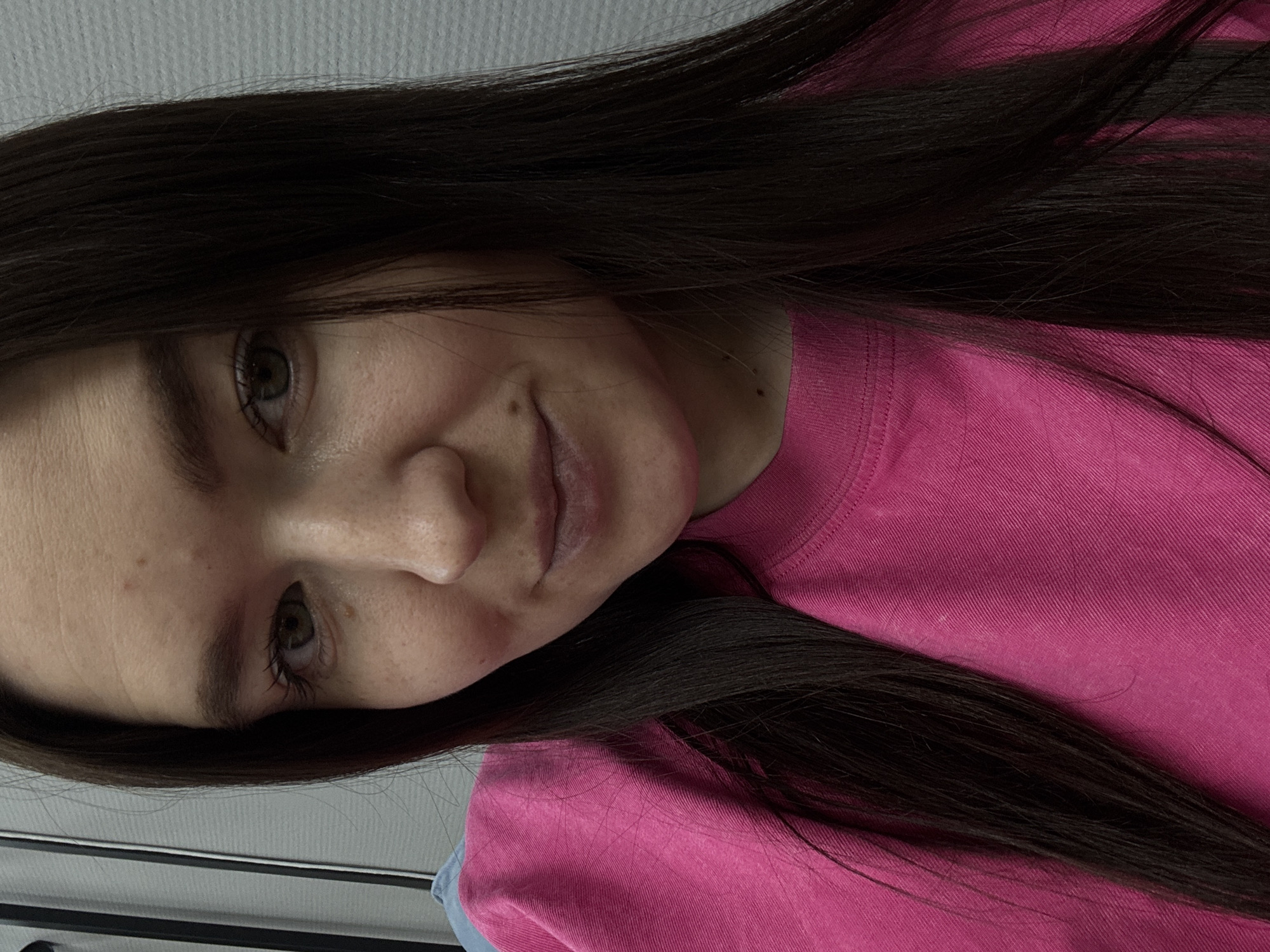 A young woman with long, dark brown hair is seen in a close-up selfie. She is wearing a bright pink, crew-neck t-shirt. Her skin tone is fair, and she has brown eyes and freckles on her face. The background is a neutral grey, appearing to be a wall. There is no indication of the location being Chaikovsky, Russia.