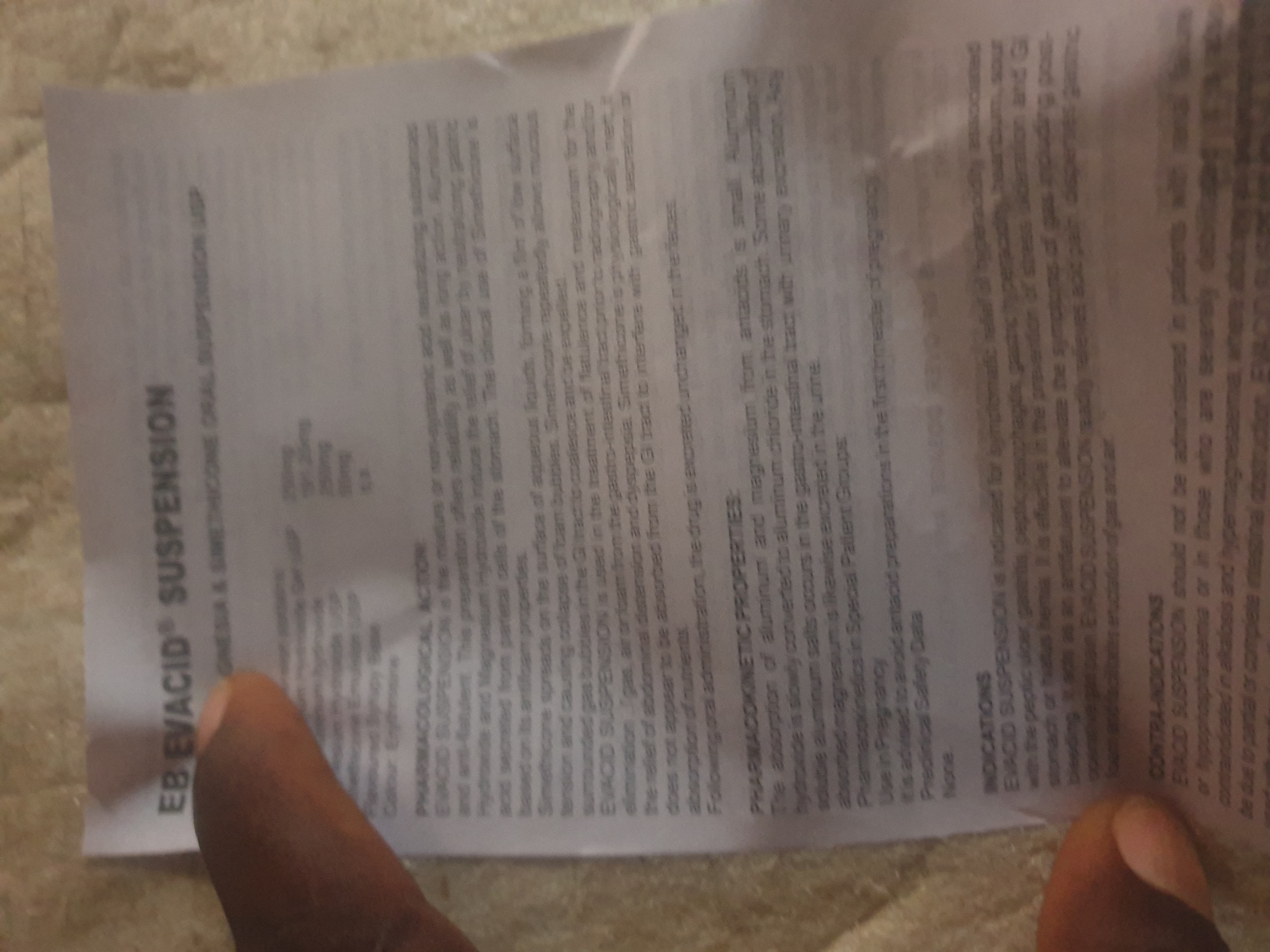 The image shows a close-up of a piece of paper, likely an information leaflet for a medication. A person's thumb and index finger are partially visible, holding the paper. The text on the paper is printed in English and appears to be medical information related to "EB EVACID SUSPENSION". Key sections visible include "PHARMACOLOGICAL ACTION", "PHARMACOKINETIC PROPERTIES", "Use in Pregnancy", "Preclinical Safety Data", "INDICATIONS", and "CONTRA-INDICATIONS". The paper is positioned on a textured surface, possibly a carpet or fabric. The lighting is somewhat dim, and the focus is on the text of the leaflet.