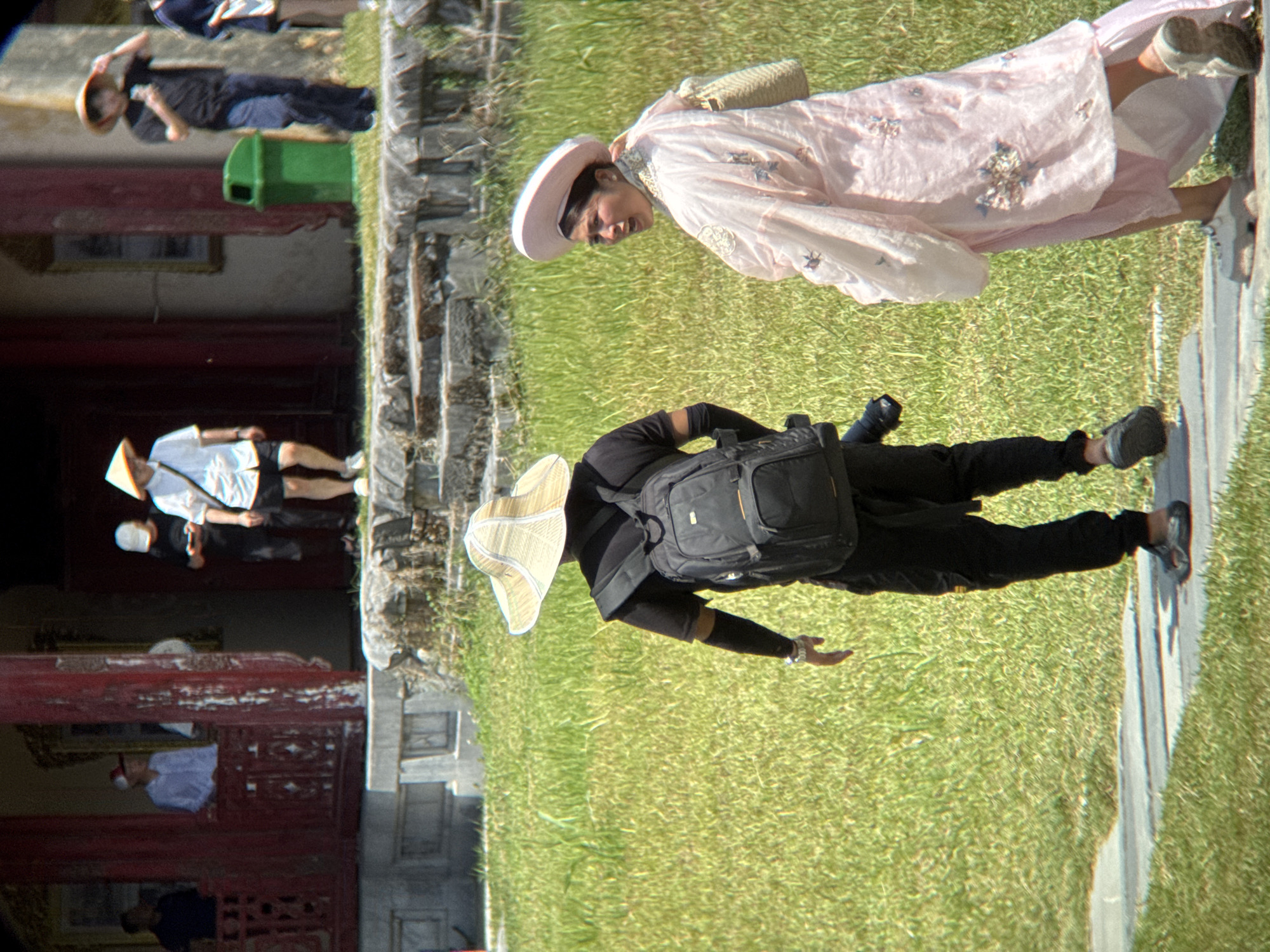 In the city of Phú Xuân Ward, Vietnam, a woman in a pink traditional dress and a white conical hat walks on a paved path in a grassy area. She carries a woven bag on her right shoulder. To her left, a person wearing a wide-brimmed straw hat, a black t-shirt, black pants, and a backpack walks away from the camera. The background features a building with red pillars and a green trash can. Further in the background, people are visible near the building entrance.