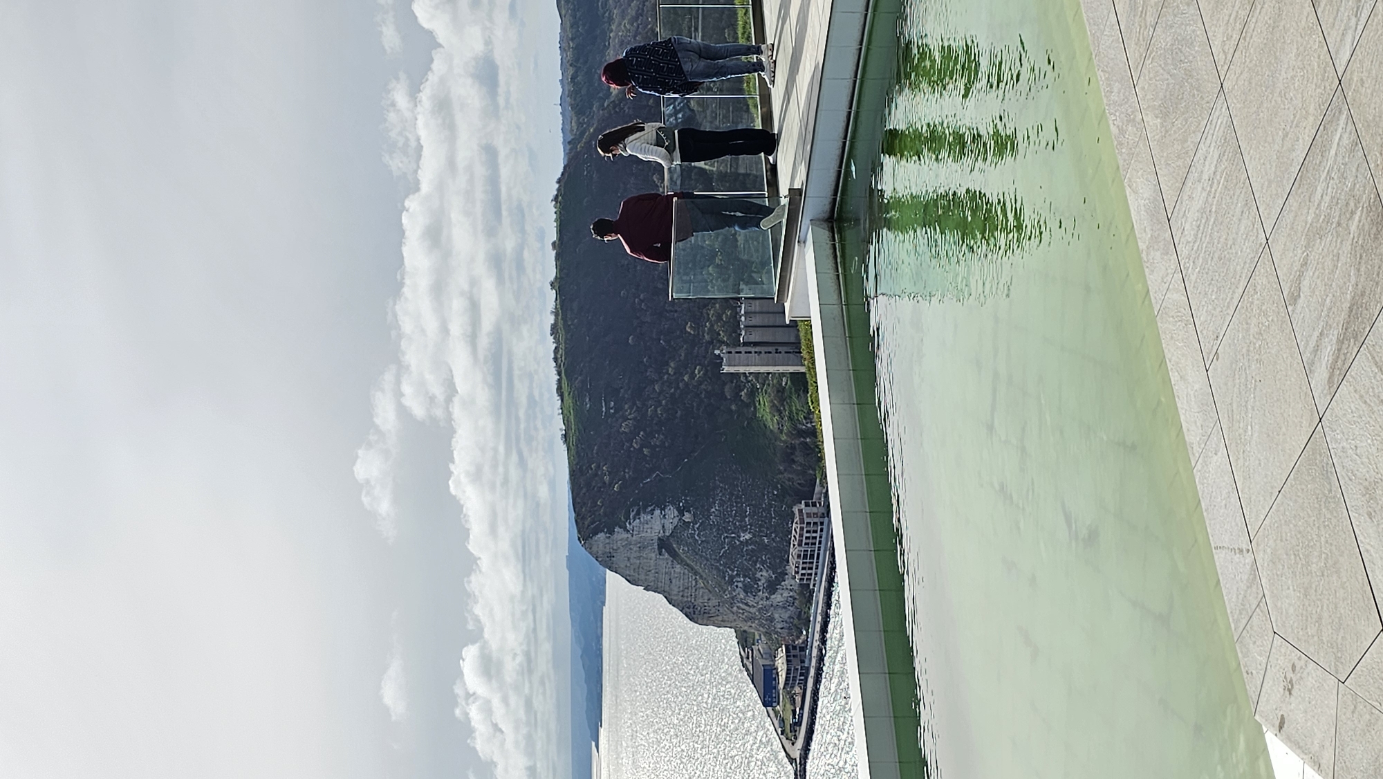 Three people stand on a building's edge overlooking a sea view in Kavarna, Bulgaria. A man in a burgundy shirt stands closest to the camera, then a woman in a white shirt, and finally another person with dark hair and a dark and light patterned shirt. They stand behind a glass barrier, with a decorative pool of water to the right and tiled decking. A large cliff with foliage and some buildings is centered behind them, with the sea visible on the left. The sky is partly cloudy.
