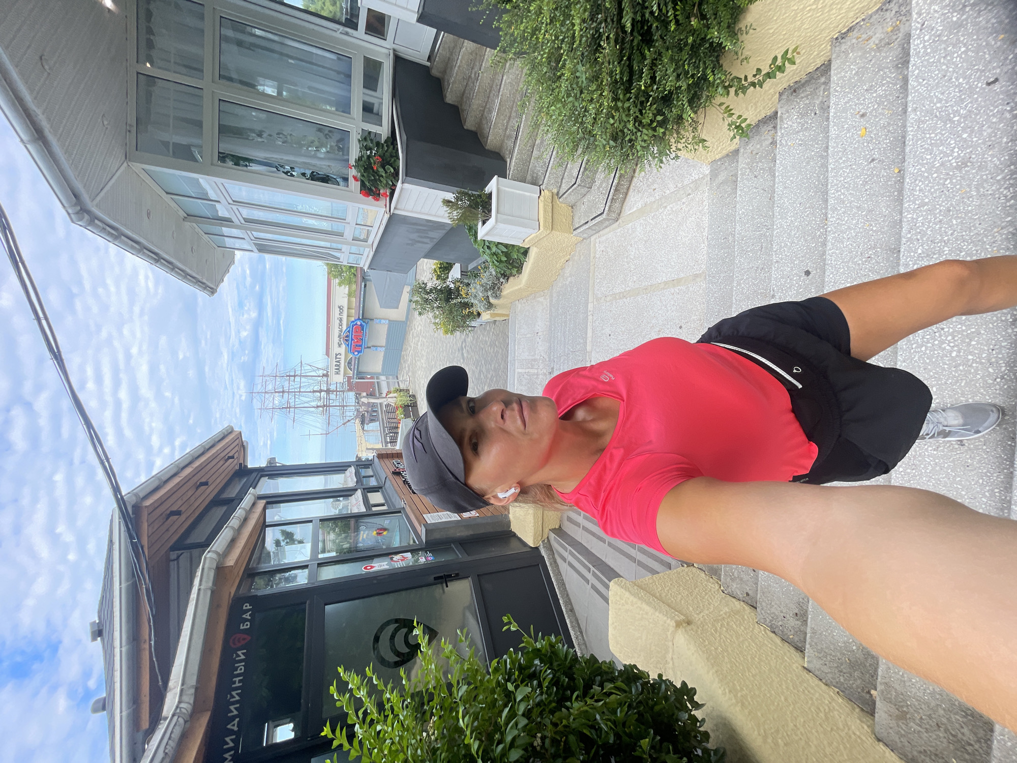 A Caucasian woman with blonde hair pulled back and wearing a pink t-shirt, black athletic shorts, a black baseball cap, and white earbuds stands on a set of outdoor stairs, taking a selfie. The stairs are flanked by buildings and lush green bushes. In the background, a large sailing ship is visible in the distance, suggesting a coastal location. Signs on the buildings indicate shops and possibly a bar. The overall scene suggests a daytime setting in a tourist area. The architecture of the buildings and the presence of a large sailing ship are characteristic of the port area in the city of Sochi, Russia, particularly near the Winter Theater and the Maritime Station, which is known for its nautical-themed attractions.