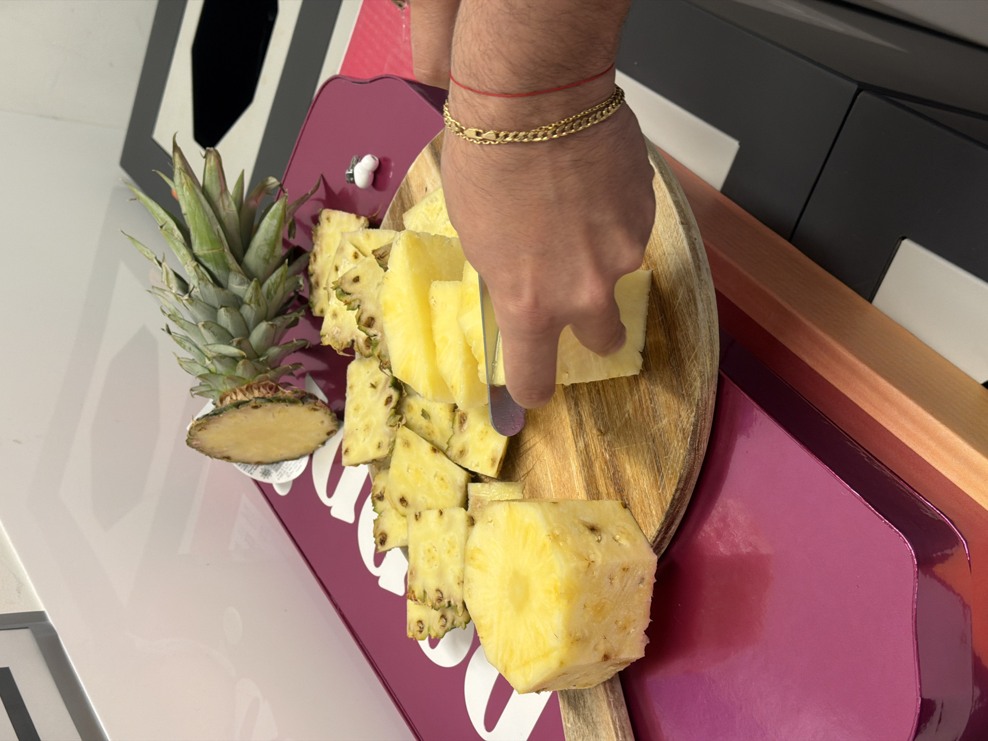 A close-up shot shows a person slicing a pineapple on a wooden cutting board. The cutting board is placed on a pink surface, which is further placed on a decorative box. The person wears a gold bracelet and a red string on their wrist. A whole pineapple and slices are visible on the left side of the cutting board. The environment includes a white surface and a black and white patterned object in the background. The scene is indoors, possibly in London, United Kingdom, but there are no obvious landmarks to confirm the location.
