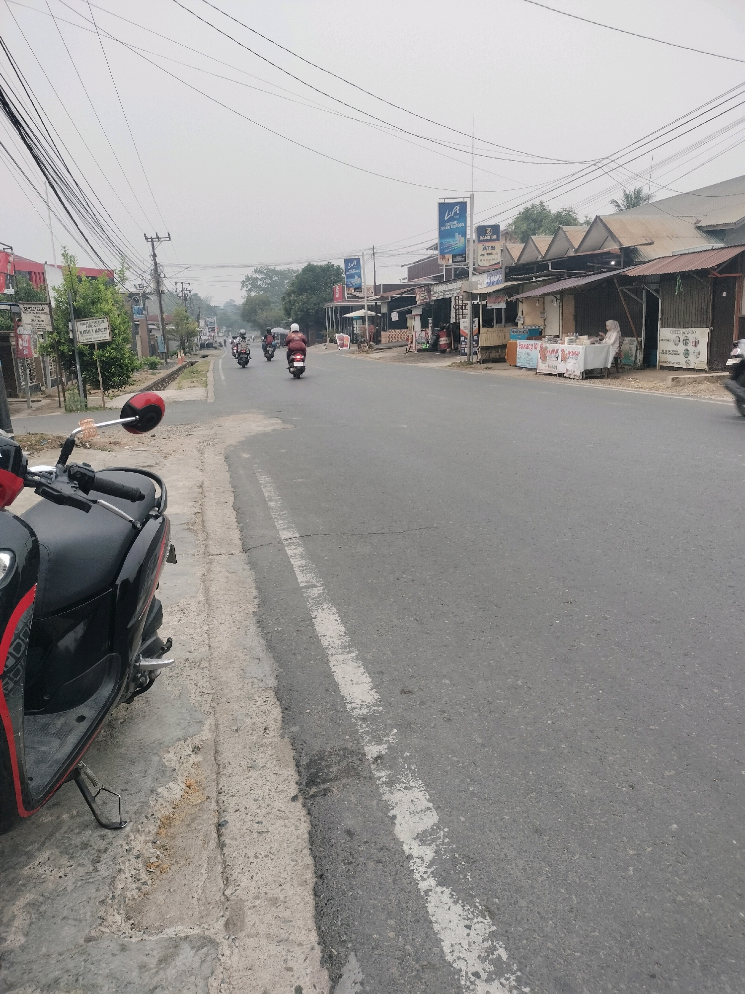 The image depicts a street scene in Pembataan, Indonesia, on an overcast day. In the foreground, a black and red scooter is parked on the side of the road, resting on its kickstand. The scooter has a red rearview mirror. The road itself is asphalt with a white painted line. To the left of the road, there is a concrete sidewalk with some debris. Along the right side of the road are various single-story structures, appearing to be small shops or stalls, with corrugated metal roofs. Some of these establishments have signage, including a blue sign with "LA ICE" and a logo, and another sign indicating "BRI ATM". In front of one of the stalls, a person wearing a hijab is seated behind a table displaying various items. Further down the road, several motorbikes are in motion, with riders wearing helmets. The background features trees and more buildings, with a network of overhead wires crisscrossing the sky. The overall atmosphere is somewhat hazy.