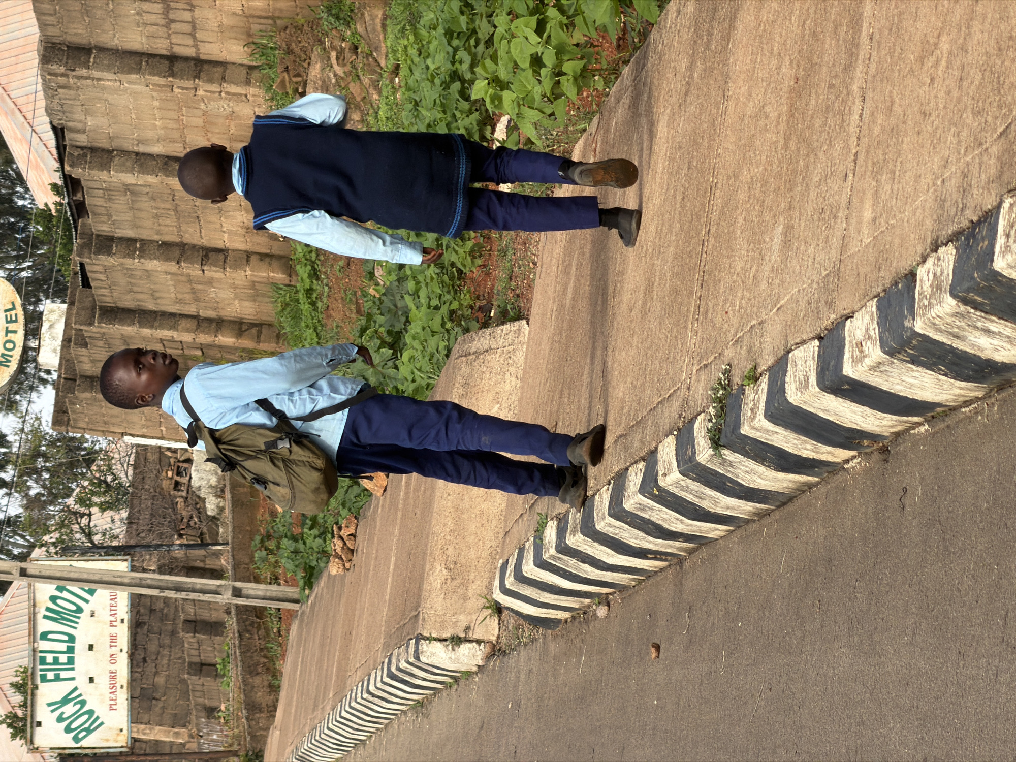 In Bukuru, Nigeria, two boys in school uniforms walk along a paved sidewalk. The boy in the foreground, wearing a light blue collared shirt and dark blue trousers, carries a khaki backpack. The boy behind him wears a light blue collared shirt and a dark blue vest over it, with dark blue trousers. They are walking away from the viewer. A road with a black and white striped curb runs alongside the sidewalk. In the background, a brick wall runs parallel to the sidewalk, with vegetation growing along its base and behind it. A sign with green lettering reading "ROCK FIELD MOTEL" and smaller text below it is visible on the left side of the image. The scene appears to be outdoors in daylight, with some sunlight casting shadows.