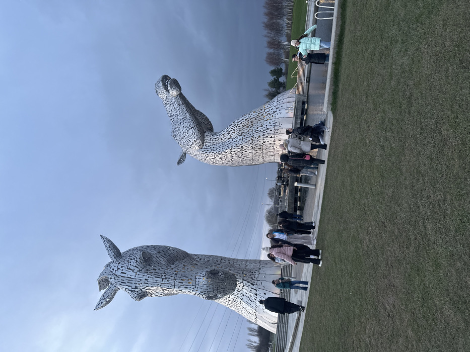 This image captures The Kelpies, two colossal equine head sculptures, situated in The Helix park in Falkirk, Scotland, United Kingdom. The scene is observed under an overcast, grey sky with low light conditions, possibly during dusk.
The two main subjects are the large, textured metallic sculptures, each composed of numerous interlocking steel plates. The sculpture on the left faces forward, while the one on the right features a curved, raised neck. Both are positioned at the edge of a dark, calm body of water.
Approximately 15 to 20 individuals are visible around the base of the sculptures. Most are standing or walking on a light-colored paved pathway that borders the water. They are dressed in casual outerwear, including jackets, coats, and some hats, suggesting cool temperatures. Actions observed include standing in small groups, individuals walking, and general observation of the sculptures. No specific interactions beyond proximity and casual conversation are discernible.
The immediate foreground consists of a sparse, dark green grassy area. In the background, power lines stretch horizontally, and bare deciduous trees are visible on the horizon. The environment is an open public park.