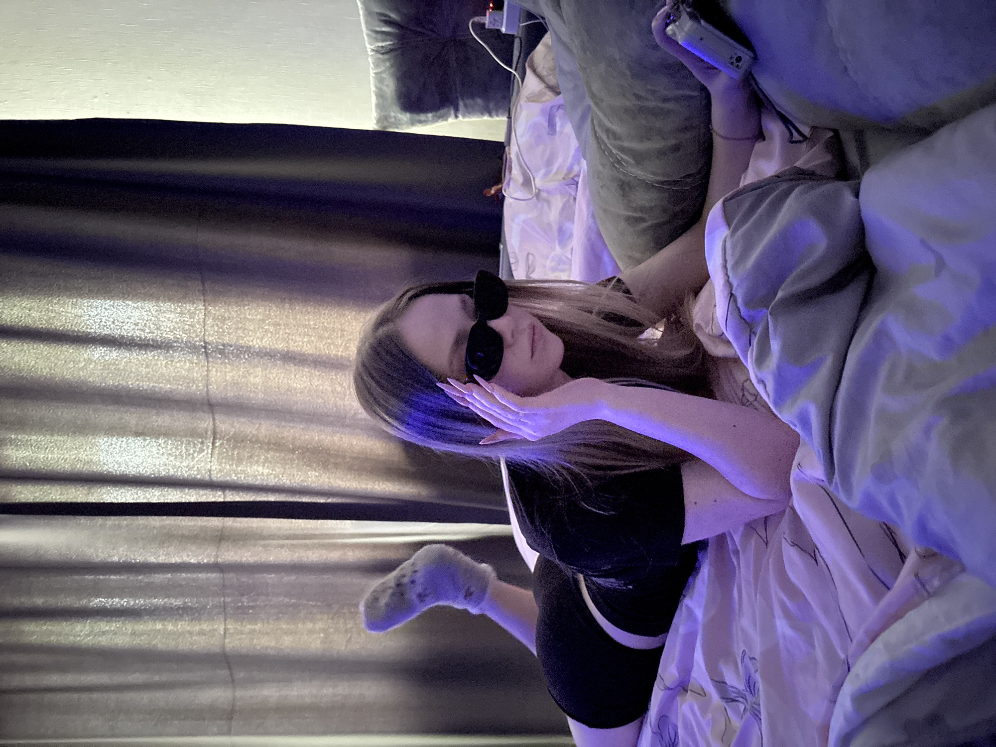 A young woman wearing sunglasses lies on a bed with floral sheets. She is wearing a black shirt and shorts and fuzzy socks on one foot. She has her hand near her face. There are curtains in the background. There is a pillow and an object that could be a remote control next to her. The image appears to be taken indoors. I do not have sufficient information to determine if this image was taken in Chaikovsky, Russia.