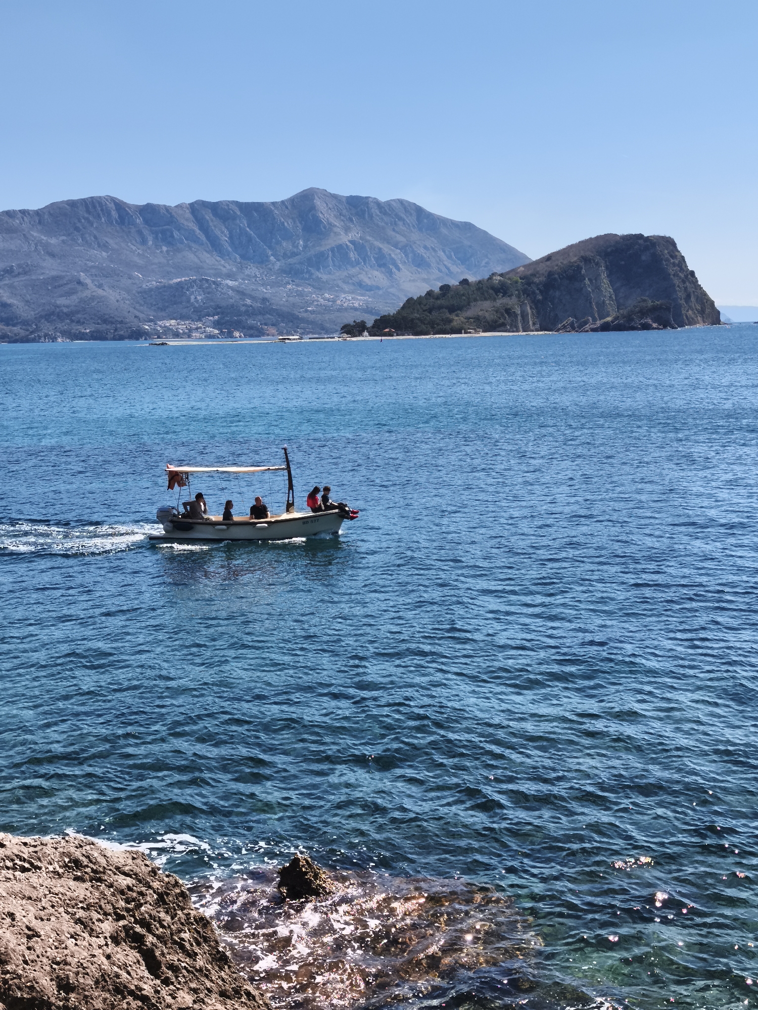 A daytime, long-shot photograph shows a small boat occupied by several individuals in the waters off the coast of Sveti Stefan in Montenegro. The boat, a light-colored vessel, is moving from left to right and has a small motor propelling it, with a wake visible behind. Several people are on board. The person closest to the back is wearing a red shirt or vest. The remaining four people are wearing casual clothing. The boat has a tan fabric awning overhead.
In the background, there's a dark, rocky island covered with vegetation, partially obscuring a sandy beach. Further in the distance, there are larger mountains. The sky is clear and blue, indicating fair weather.
In the foreground, there are jagged rocks in the water, indicating that the photograph was likely taken from a coastal area. The water is a deep turquoise color.