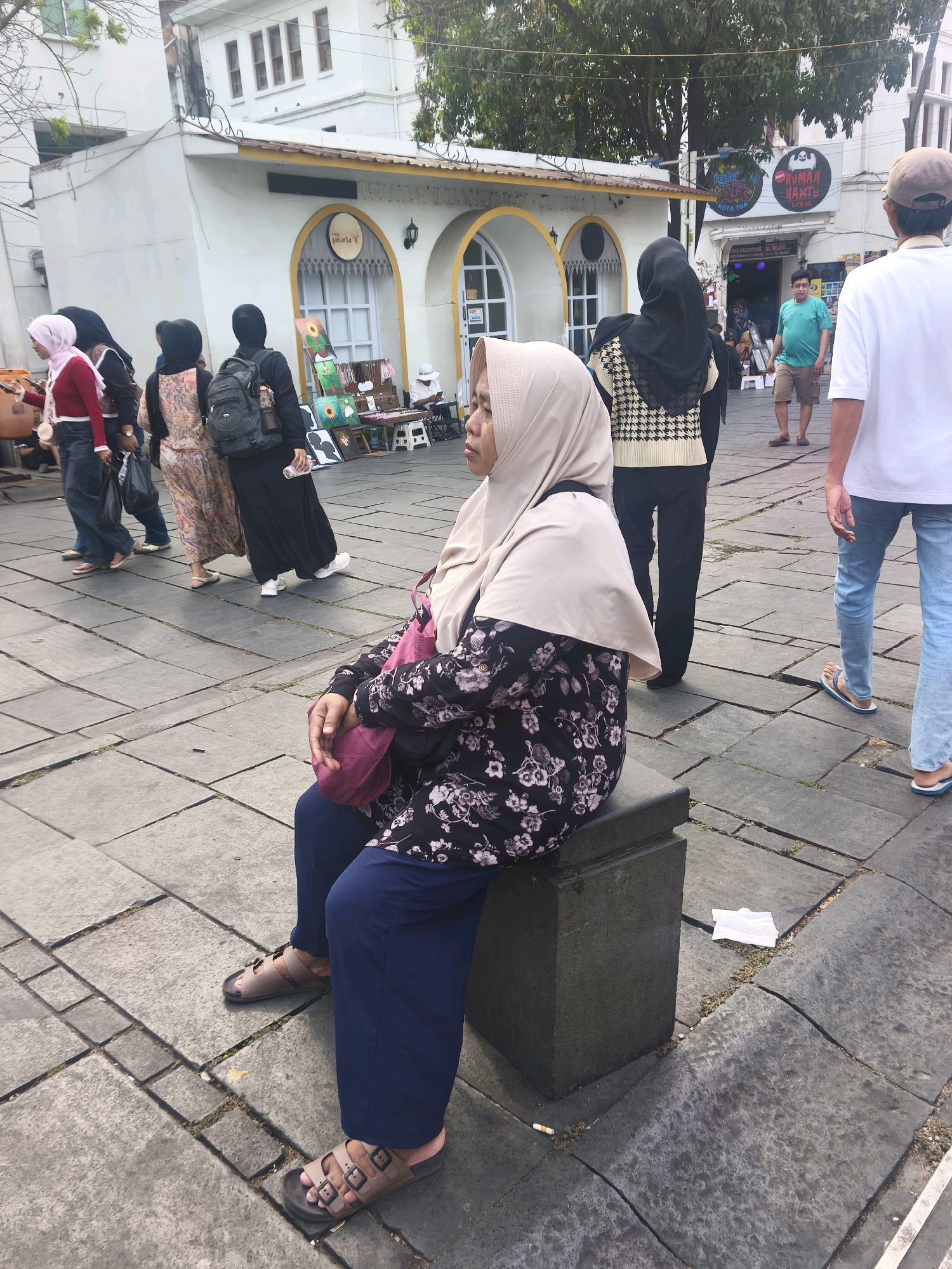 In a public square in West Jakarta, Indonesia, several individuals are walking or sitting on stone paving. In the foreground, a woman wearing a light-colored hijab and a floral-patterned blouse sits on a dark stone block, her legs crossed and her hands resting in her lap. She carries a pink bag over her shoulder. Behind her, other people are seen in various states of movement. To the left, a group of women in hijabs are walking, one in a red jacket and jeans, another in a floral dress, and another in a black abaya with a backpack. Further in the background, a man in a turquoise shirt and shorts stands while another man in a white t-shirt and jeans walks away. Buildings with arched windows and awnings line the background, some displaying signs. The environment appears to be an outdoor marketplace or pedestrian area.