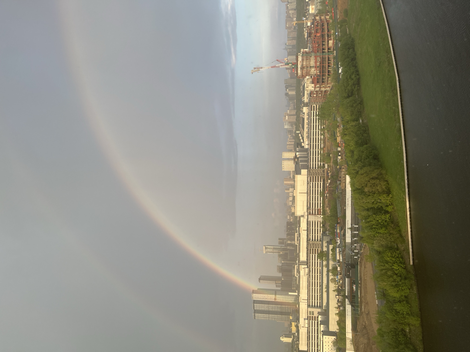 The image depicts a partial rainbow over the city of Khimki, Russia. The rainbow is seen on the left side of the frame, arching over the urban landscape. The cityscape consists of numerous high-rise buildings, construction sites with cranes, green spaces, and a body of water on the right edge. The buildings vary in height and architectural style, indicating a mix of residential and commercial properties. Construction is ongoing, evidenced by the presence of cranes and partially completed structures. A strip of green space, likely a park or greenway, runs along the edge of the water. The overall impression is of a modern, developing city with active construction and urban green spaces.