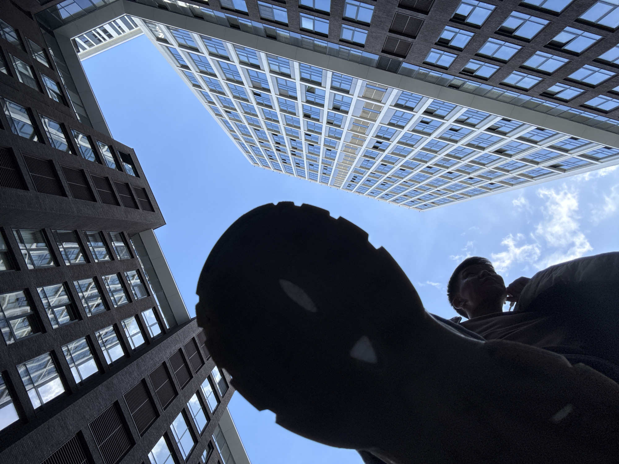 The image presents an upward-looking perspective within a city environment. Two high-rise buildings, likely office or apartment complexes, frame the sky. The building on the left appears to be of darker construction with a grid of windows, whereas the building on the right is lighter, possibly constructed of white concrete or light-colored materials. The sky is visible between the buildings, displaying a clear blue with some scattered clouds. In the foreground, there is a person looking up, with a large black silhouette, which appears to be the sole of a shoe. The lighting indicates a sunny day, casting the silhouette against the sky. Based on the architectural style of the buildings, it is likely that this photograph was taken in the city of Moscow, Russia.