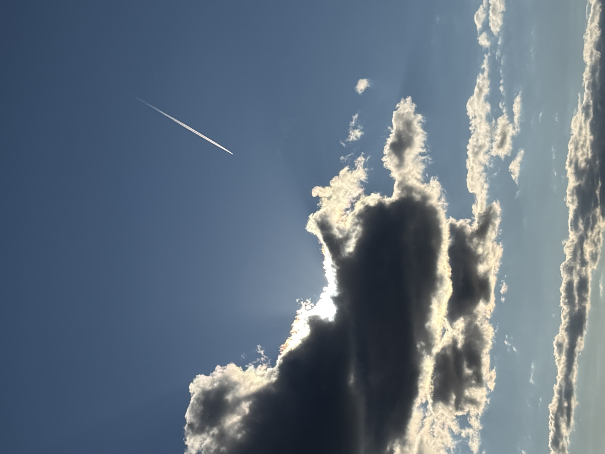 An airplane contrail streaks across a clear blue sky above Chaikovsky, Russia. The sun is partially obscured by a large, dark cloud, casting rays of light through the gaps. Additional scattered clouds are visible in the lower portion of the image, some illuminated by the sun. The scene appears to be a natural landscape, with no people or man-made structures other than the airplane's contrail.