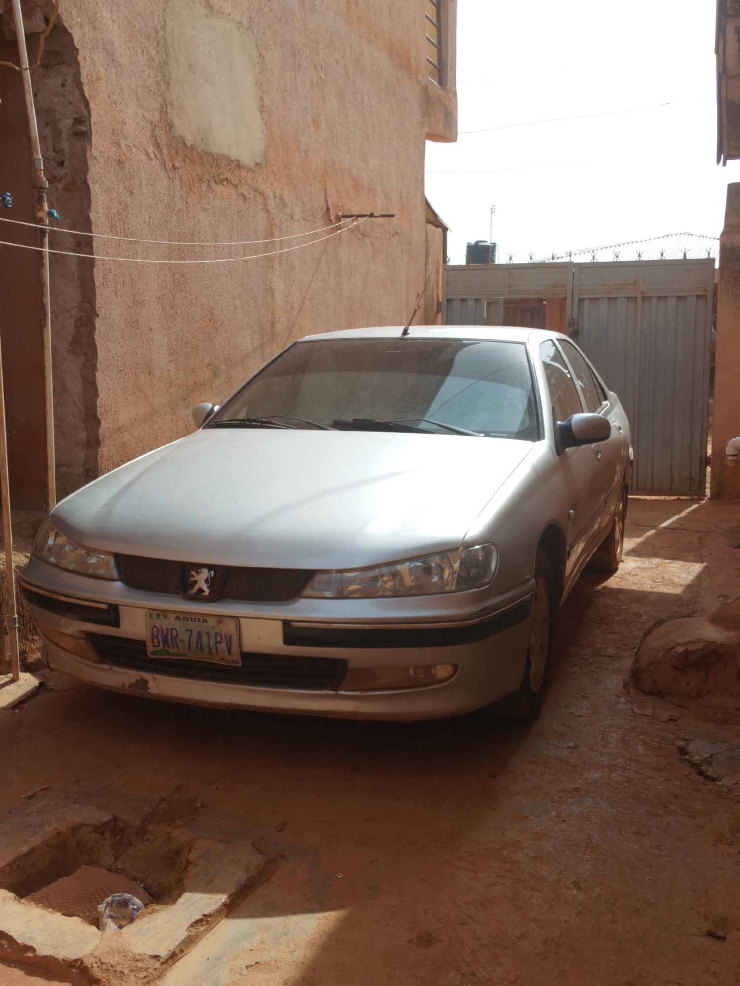 A silver Peugeot 406 sedan is prominently parked in a narrow, unpaved alleyway or driveway. The car is oriented slightly towards the left, displaying its front and left side. Its front license plate is clearly visible, reading "BWR-741PV" with "ABUJA" printed above it, accompanied by a small Nigerian flag symbol. The distinctive Peugeot lion emblem is also visible on the grille.
The car is situated between two rough, earthen-toned walls, likely part of residential or commercial buildings. These walls show signs of wear and age, with patches of crumbling or discolored plaster. On the left wall, some exposed piping is visible, and two thin white wires are strung horizontally across the alley, possibly for laundry. The ground is unpaved, consisting of dirt and dried mud, and features a noticeable open drain or channel in the foreground containing a discarded plastic bottle.
In the background, a tall, light-colored metal gate with corrugated panels and barbed wire along its top edge spans the width of the alley, suggesting an entrance to another property. Behind the gate, a black water tank mounted on a stand is partially visible. The sky above appears bright and clear, indicative of sunny weather, with strong shadows cast by the car and surrounding structures suggesting it is mid-day. There are no people visible in the scene, and the car is stationary. The setting, including the architecture, unpaved ground, and visible infrastructure like the water tank, points to a residential or semi-urban environment in Nigeria.