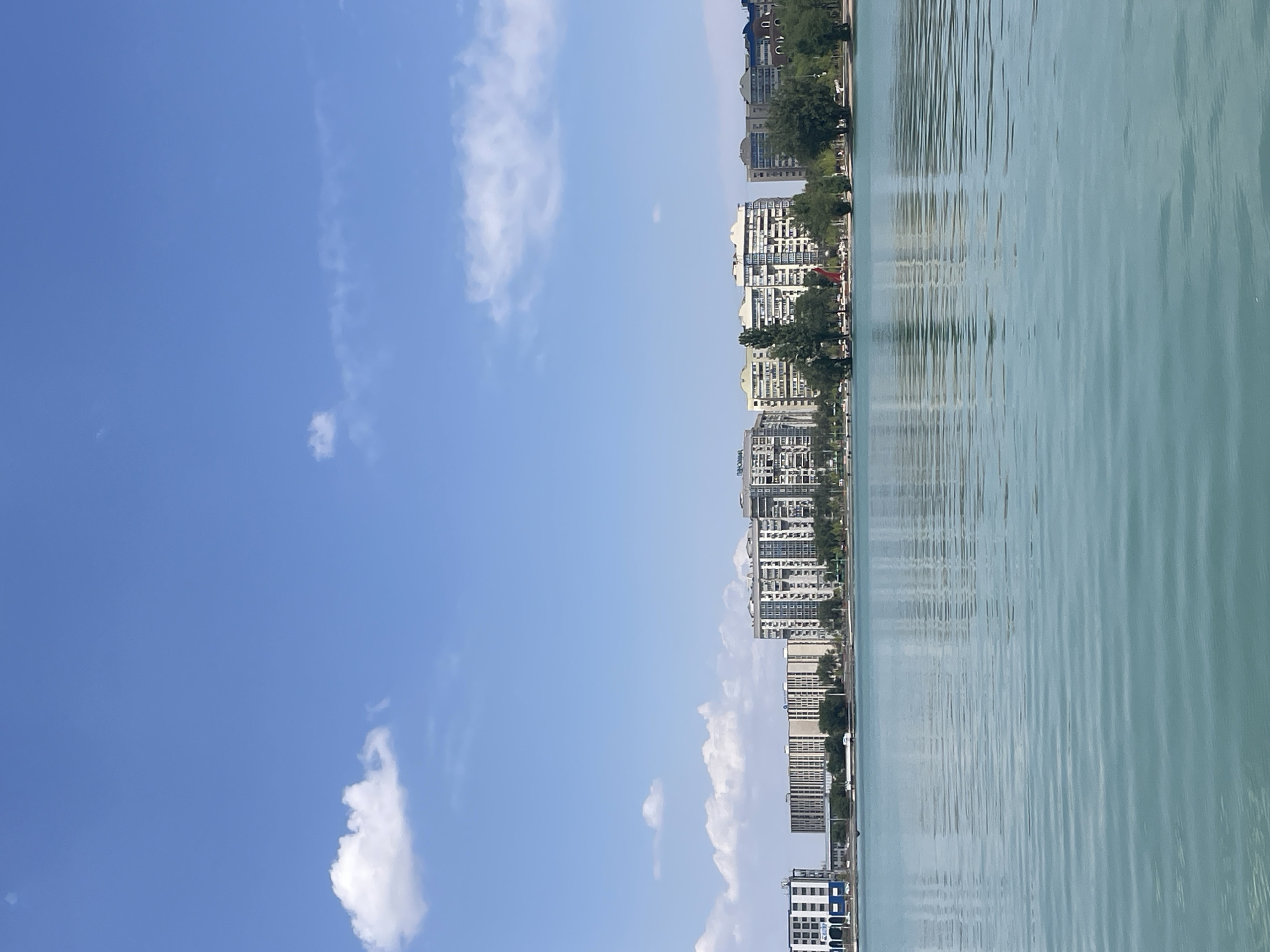 The image depicts a serene view of the Big Almaty Lake in Almaty, Kazakhstan. The foreground is dominated by the tranquil, turquoise waters of the lake, with gentle ripples creating a textured surface. The water reflects the clear blue sky and the buildings lining the shore.
In the background, a line of modern apartment buildings stretches across the horizon. These buildings are predominantly light-colored, with many windows and balconies. Some of the buildings have varying architectural styles, suggesting they were built at different times. Lush green trees and vegetation are interspersed between the buildings, adding a touch of nature to the urban landscape. The sky is a bright, clear blue, with a few wispy white clouds scattered across it, indicative of a sunny day. The overall impression is one of urban development coexisting harmoniously with a beautiful natural body of water.