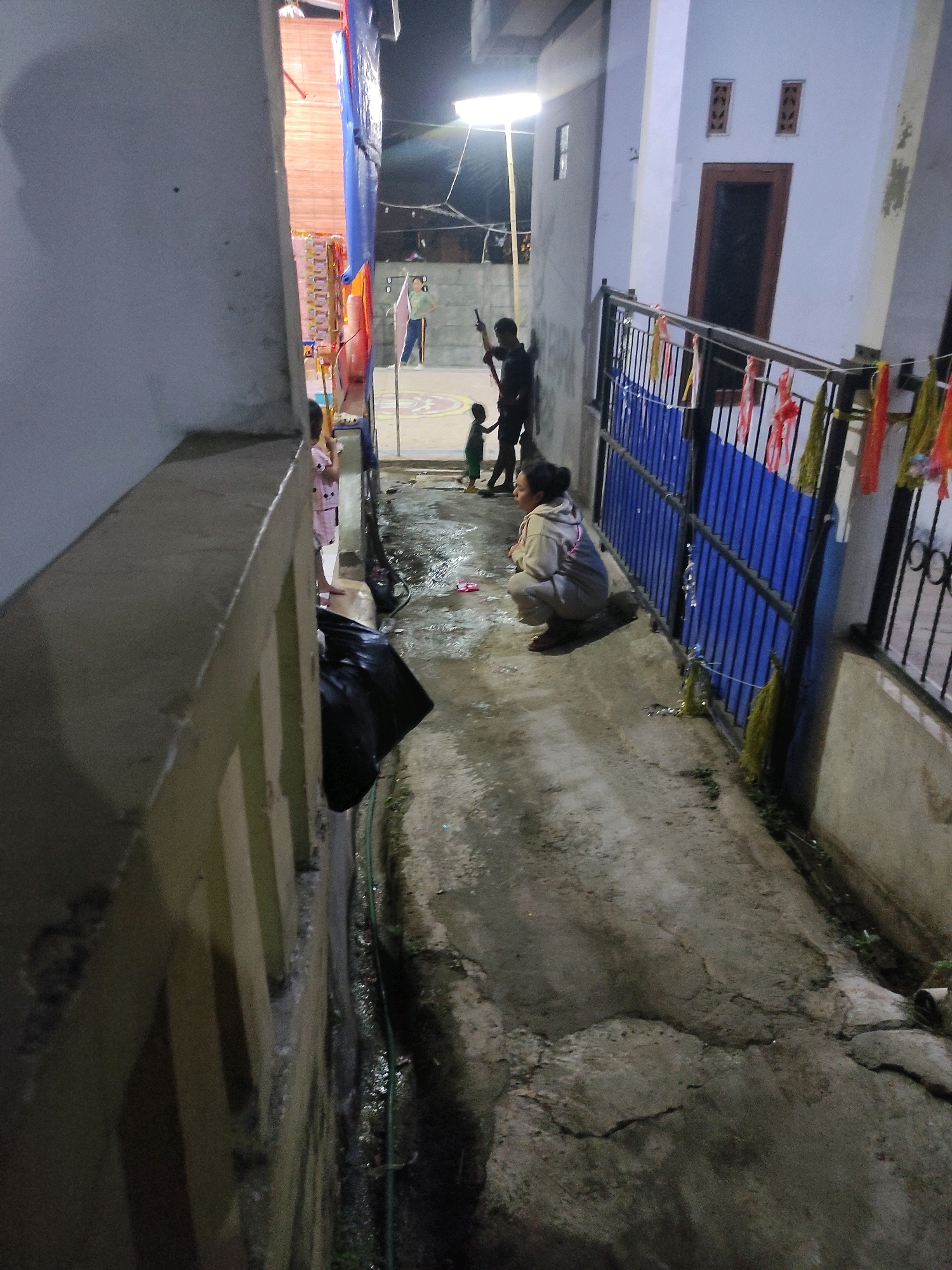 The image shows an outdoor scene at night, likely in Padalarang, Indonesia, featuring a narrow pathway between buildings. In the foreground on the left, a light-colored wall with vertical elements is visible. The pathway itself is made of cracked concrete and shows signs of moisture and some greenery growing along the edges.
Several people are present in the scene. In the middle ground, a woman is crouching on the pathway, facing left, wearing a light-colored hoodie and pants. To her left, a young child in a green shirt stands near a person whose silhouette is visible, holding what appears to be a stick. Further back, another person, possibly an adult, is also visible in silhouette, holding a stick.
On the right side of the pathway, there is a black metal fence with blue and red decorative elements hanging from it. Behind the fence, a white building is partially visible, with a brown-framed window and two small decorative openings. Another building with a blue tarp is also visible in the background. A bright light source is illuminating the scene from above. A black plastic bag is placed on the ground near the left wall.