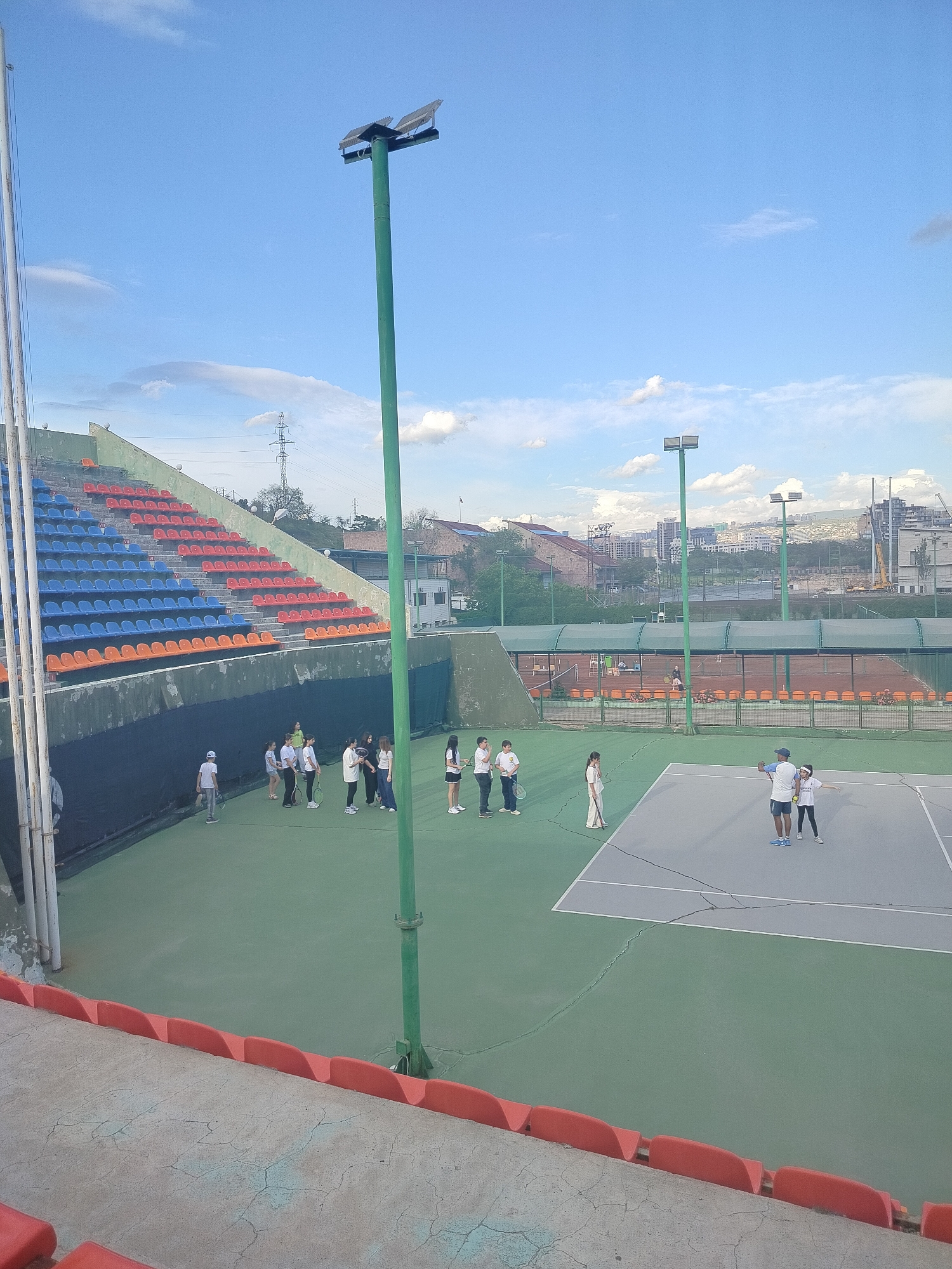 The image depicts a tennis court with an audience stand in Yerevan, Armenia. The stand has rows of red, blue, and orange seats. A group of about ten people stand on the court, appearing to be receiving instruction from a coach. Another court is visible in the background, with a canopy and additional players. Beyond the tennis courts, the urban landscape of Yerevan is visible, with buildings and a construction crane. The sky is blue with scattered clouds. Tall, green lampposts are positioned around the tennis court.