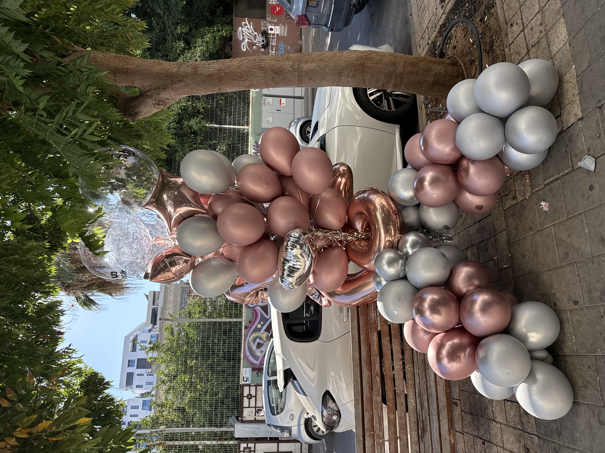 In the city of Tel-Aviv, Israel, on a sunny day, a bouquet of rose gold and silver balloons is displayed on a wooden bench. The balloons, including clear ones with confetti inside, are arranged in a large cluster and are attached to a number foil balloons, one of which appears to be the number '2'. Behind the balloons and bench, a white car is parked, and a tree with green foliage provides shade. In the background, there are buildings, a fence, and graffiti on a wall. The ground is paved with interlocking bricks. There are no people visible in the image.
