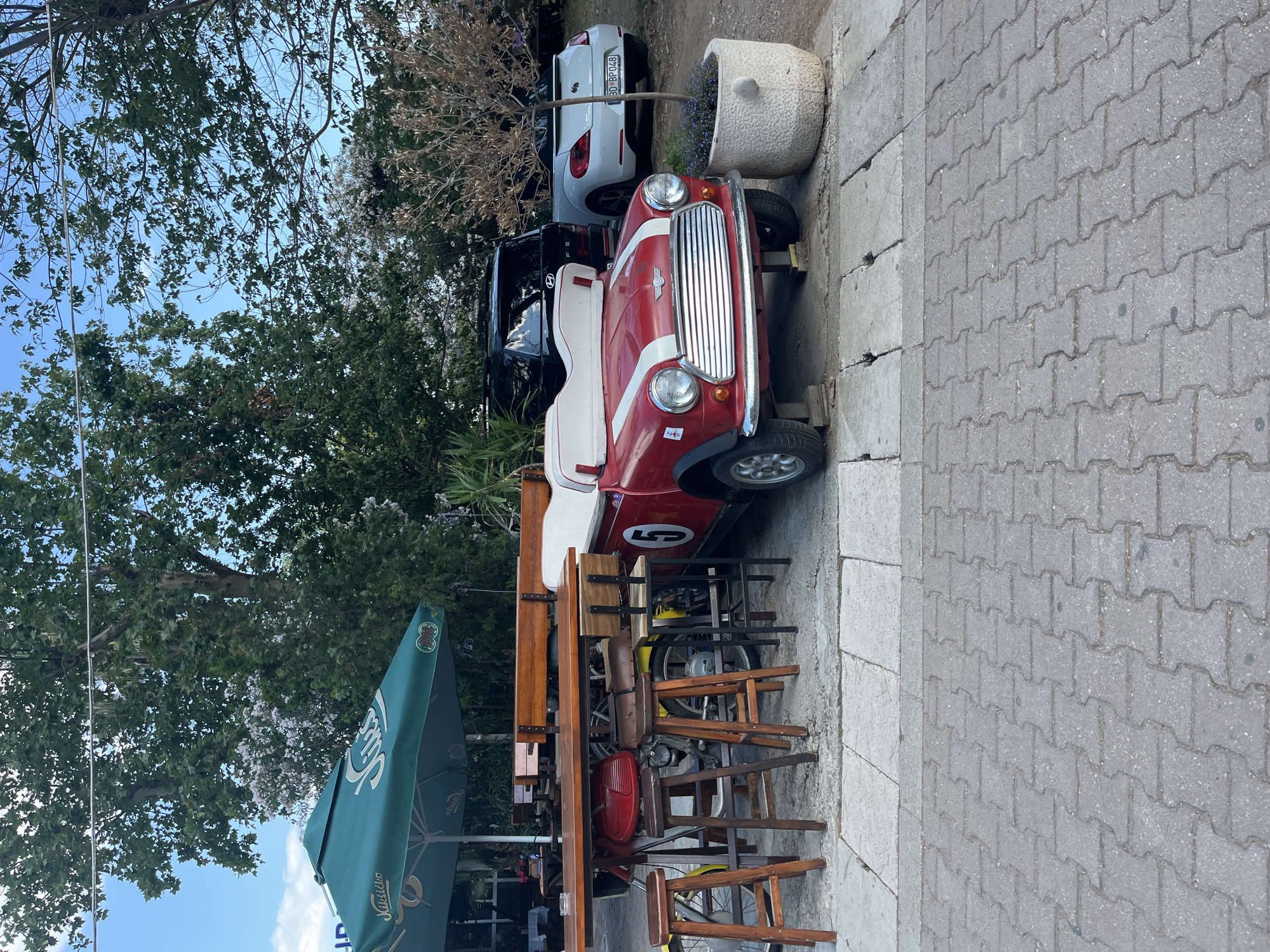 This media file depicts an outdoor scene, likely a cafe or restaurant seating area, in Budva, Montenegro. The focal point is a distinctive vintage red and white mini-car, possibly a golf cart or a modified classic car, parked on a paved walkway. The car features a chrome grille, circular headlights, and the number "5" prominently displayed on its side.
Behind the mini-car, a modern white car is visible, suggesting a juxtaposition of old and new. A large, decorative pot filled with plants sits next to the white car. The setting is outdoors, with lush green trees and foliage framing the scene, indicating a pleasant, possibly warm, time of day with clear skies. Sunlight casts shadows, suggesting it's daytime.
In the foreground, a wooden table and several chairs are arranged, implying a space for dining or relaxation. A green umbrella, branded with text that appears to be "JULES" and "MIRANO", is partially visible, further reinforcing the idea of an establishment. The paved walkway is made of interlocking concrete pavers.
There are no people visible in the frame. The overall mood is relaxed and perhaps a bit quirky due to the unique vehicle. The scene appears to be set in a lively tourist area, given the presence of cafes and the distinctive car as a potential attraction. The time of day is likely afternoon based on the lighting.