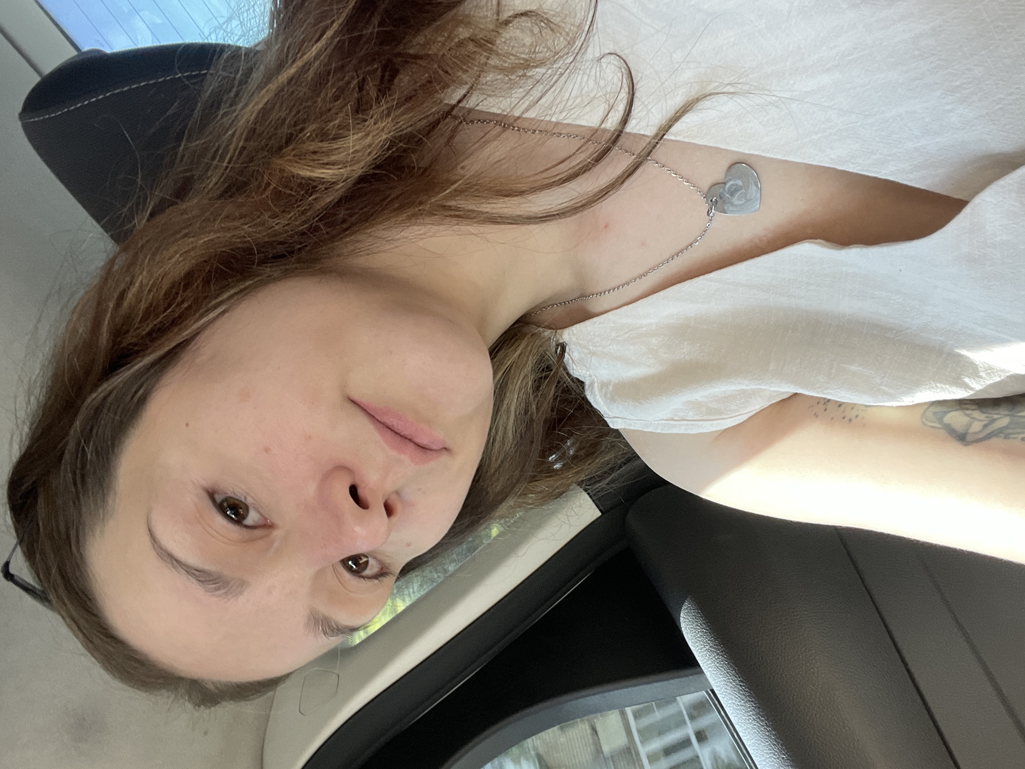 The image is a close-up selfie of a young woman taken inside a vehicle. The woman has light brown hair and brown eyes. She is wearing a white top and a silver heart-shaped pendant necklace. The background shows the interior of a car, with a headrest and a window visible. Sunlight is casting shadows across the scene. There is no indication of the specific location within Stavropol, Russia.