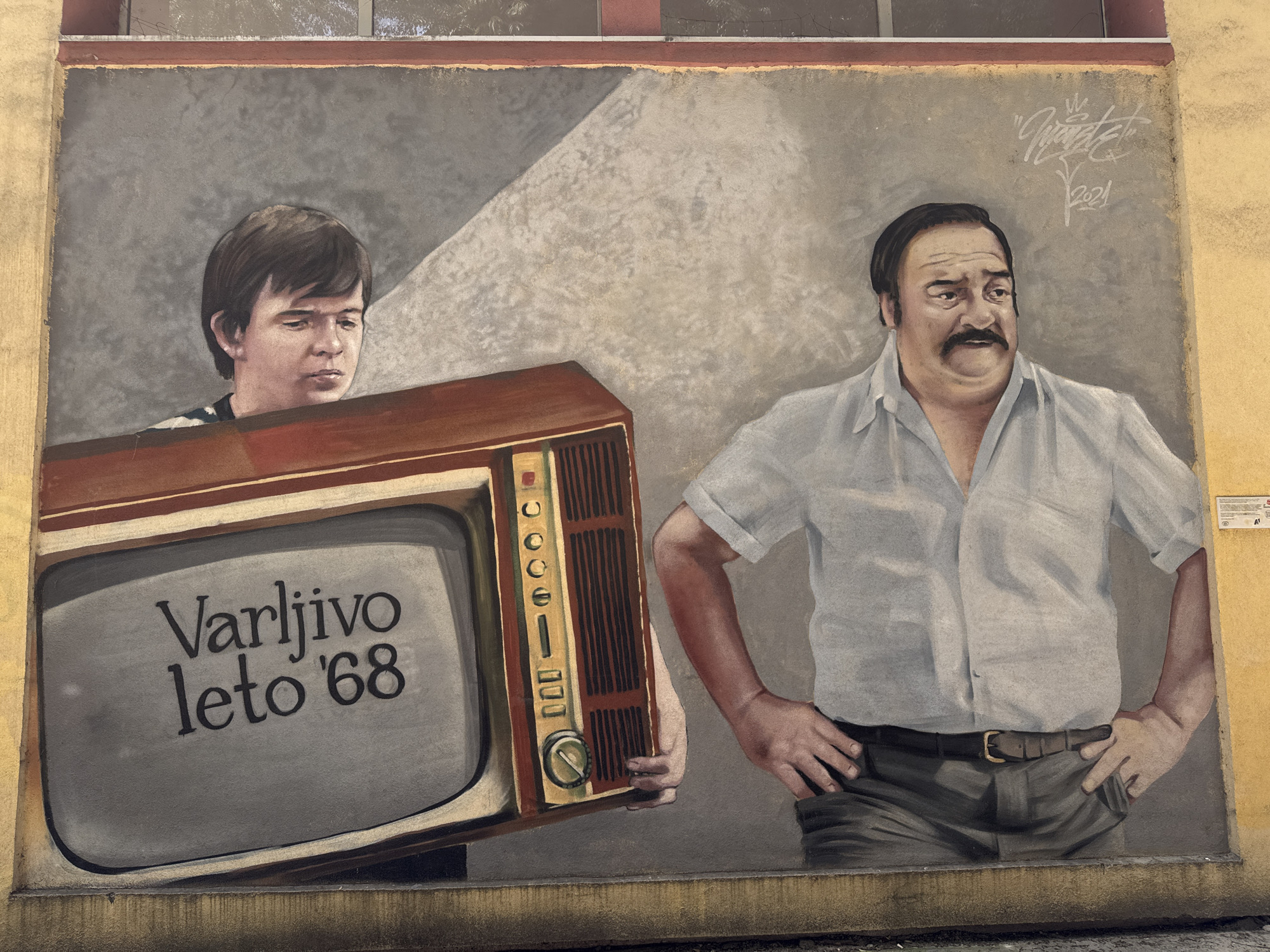 The image shows a mural on a building wall in Novi Sad, Serbia, depicting characters from the Yugoslav film "Varljivo leto '68" (Treacherous Summer of '68). A young man carries an old television with the film's title displayed on the screen. An older man, likely the character of the father, stands beside him with his hands on his hips. The mural's style suggests it's a recent artwork. The top of the mural reaches the window of a brick building and is at street level.