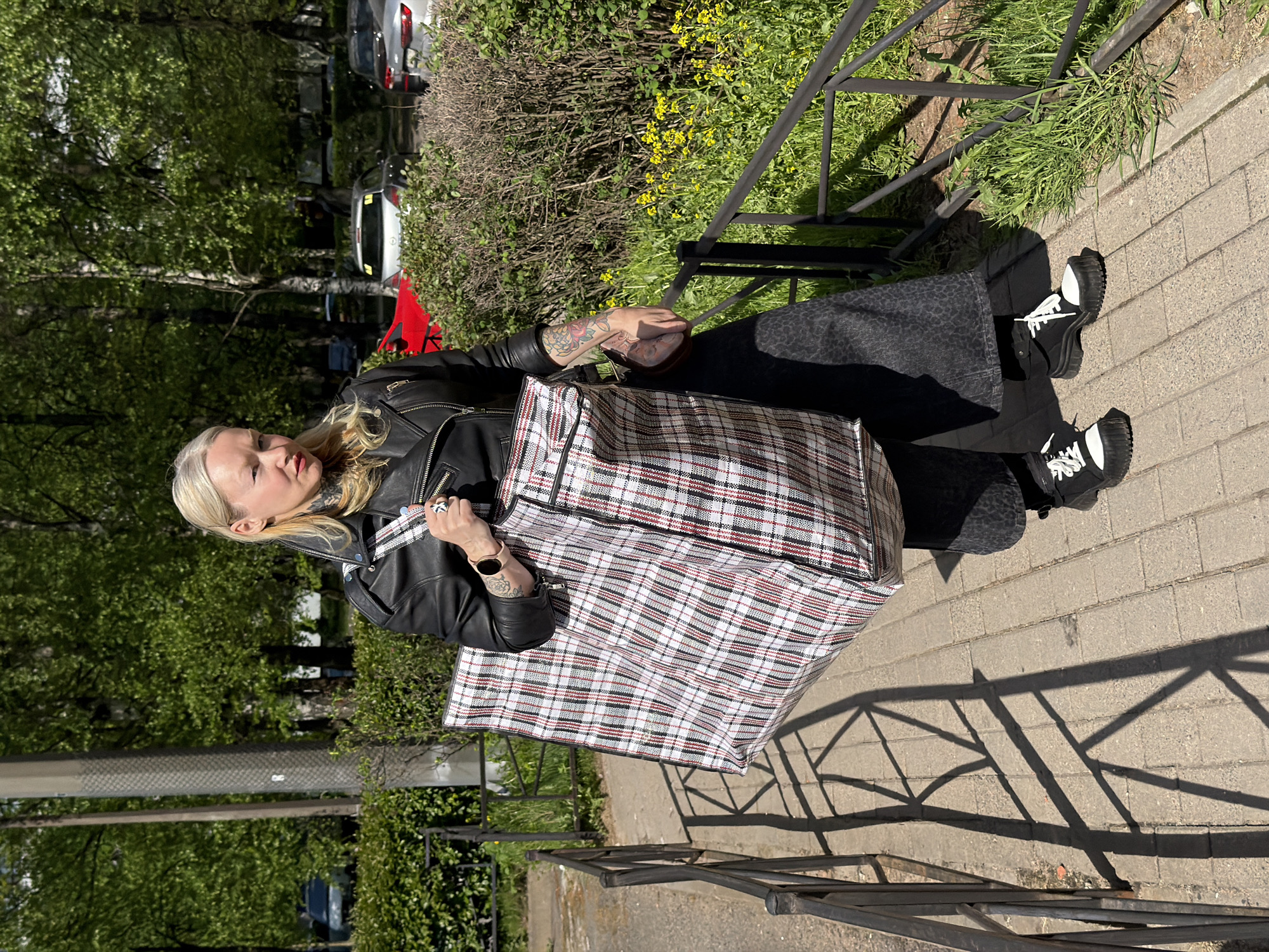 A woman stands on a paved sidewalk near a metal fence, holding a large, plaid bag. She wears a black leather jacket, patterned black pants, and black and white shoes. Tattoos are visible on her arms and neck. The background shows trees and bushes, with cars parked along the street. The scene is likely in Saint Petersburg, Russia, based on the style of architecture and urban environment.