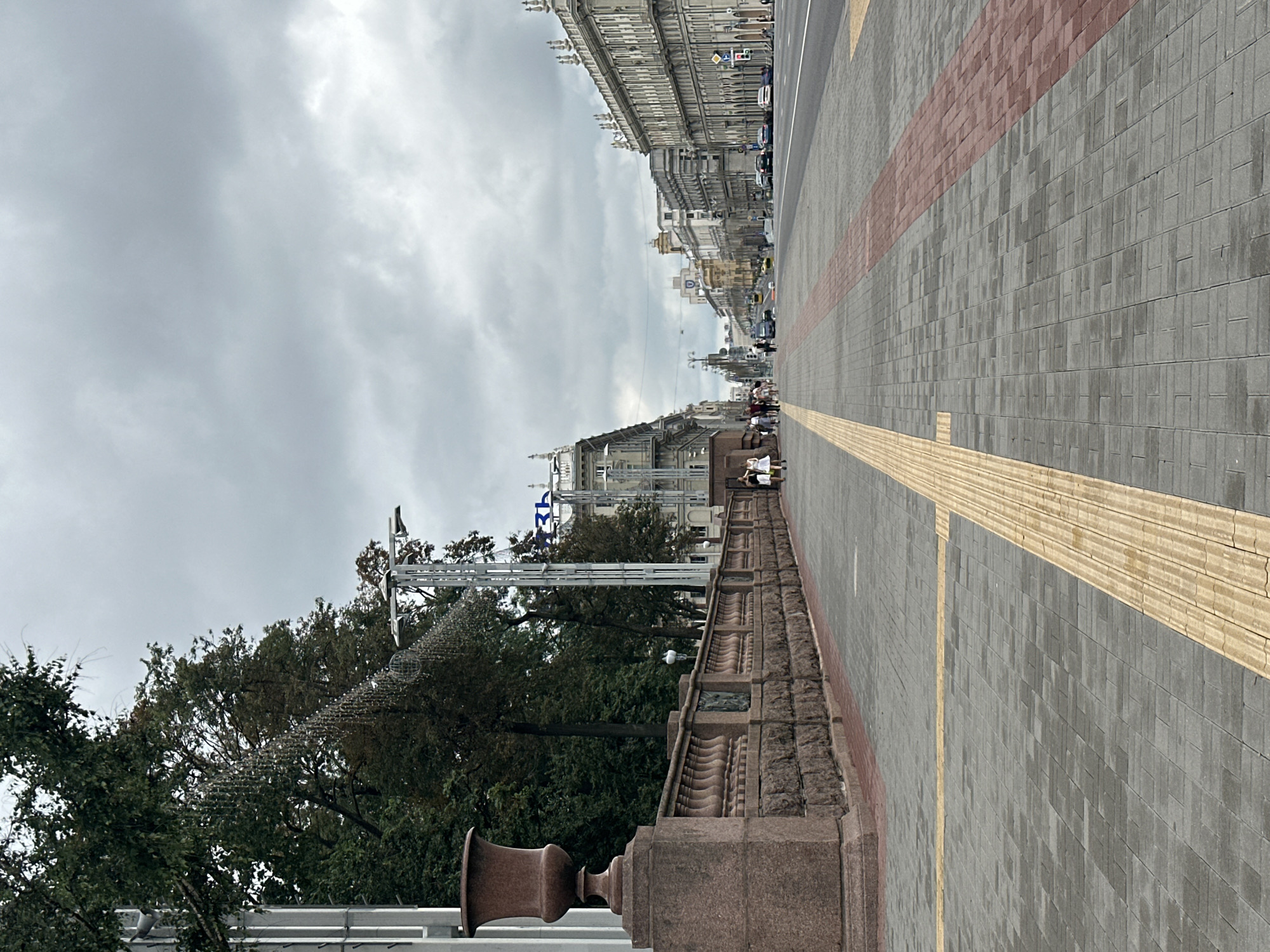 This is a medium-shot, eye-level, vertical, outdoor, daytime, slightly overcast photograph taken on Independence Avenue (Praspyekt Nyezalyezhnastsi) in Minsk, Belarus. The perspective looks down the wide avenue, with a pedestrian walkway on the left and a multi-lane road on the right. The walkway is paved with grey rectangular blocks, with a tactile paving strip of yellow blocks running along the edge closest to the street. A red brick-patterned strip runs parallel to the street further down the pedestrian area.
On the left side of the walkway, a large stone balustrade with ornate carvings runs along the edge of a raised embankment. A massive, dark reddish-brown stone urn sits atop a stone pedestal at the start of the balustrade in the foreground. Behind the balustrade, lush green trees form a dense backdrop. Some decorative string lights are visible hanging from poles and trees. A tall, modern silver structure with a globe-like ornament is positioned further down the avenue, near the trees.
Several pedestrians, mostly women in casual clothing, are walking along the sidewalk in small groups. Further down the avenue, cars are driving on the road, and more pedestrians are visible on the sidewalk. The architecture on both sides of the avenue consists of grand, multi-story buildings with classical and Soviet-era architectural elements, featuring elaborate facades, columns, and decorative cornices. The sky is filled with heavy, grey clouds, suggesting a dull or impending weather. The overall scene depicts a wide urban thoroughfare with a mix of historical and modern elements, typical of a major European city.
