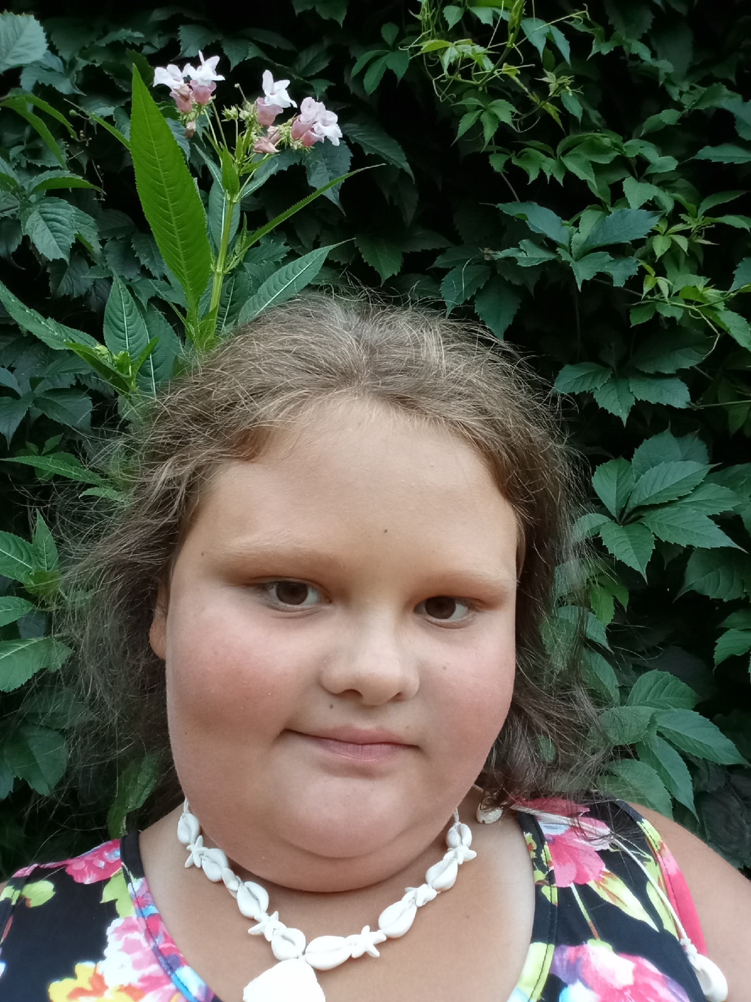 A young Caucasian girl with light brown hair and brown eyes is pictured from the chest up, facing the camera. She is wearing a floral print dress and a white shell necklace. She is standing outdoors in front of a dense wall of green foliage and some flowering plants. The image was taken in Pavlovsky Posad, Russia.