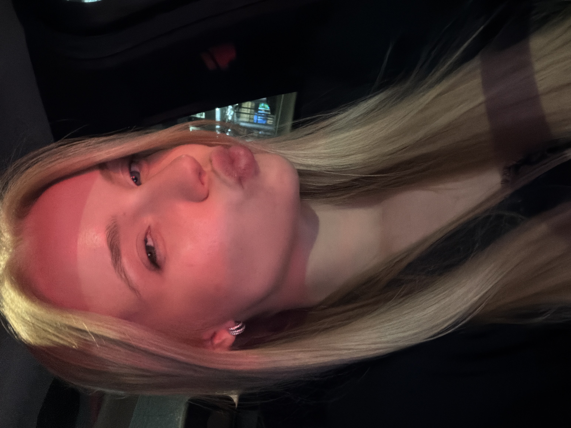 A young woman with blonde hair and fair skin poses for a selfie in a dark environment. She is making a kissing face with her lips pursed. She is wearing earrings. Reflections of lights from a building can be seen in the background. The location is in a city, Unknown, Unknown.