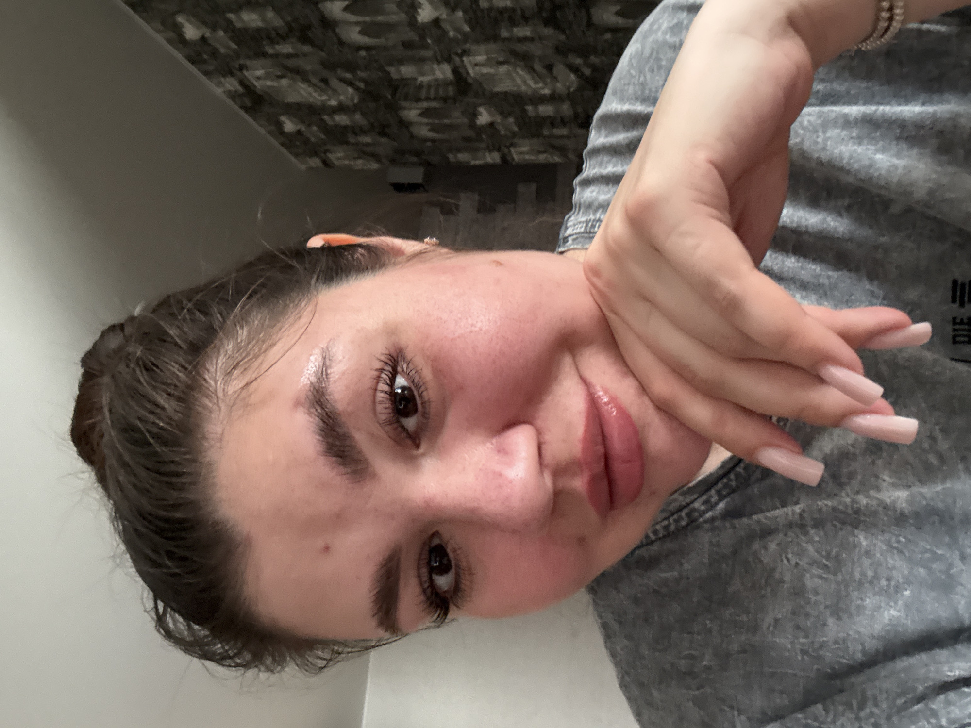 A young woman poses for a close-up, angled selfie. She rests her chin on the fingers of her right hand, which is adorned with long, pink, manicured nails. She has brown hair pulled back, brown eyes with prominent eyelashes, and appears to be wearing lip gloss. A bracelet is visible on her wrist. She wears a gray shirt. The background is a blurred mix of wall and ceiling. The image's content does not suggest a specific location, especially not Votkinsk, Russia.