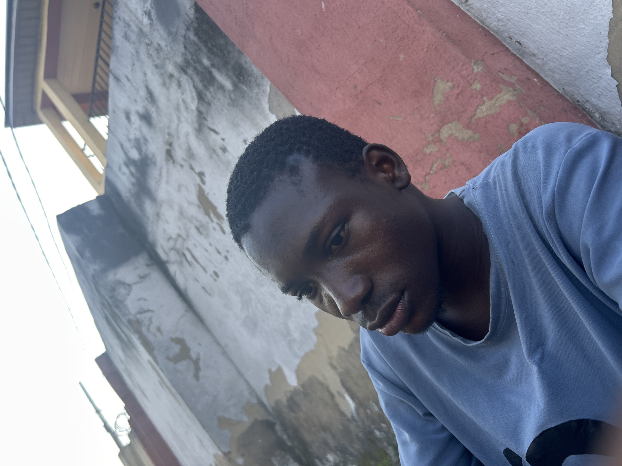 The image is a close-up, low-angle shot of a young Black man with dark skin and short, dark hair, wearing a light blue t-shirt. He is looking off to his left, with his head slightly tilted. The man has a small goatee. The background consists of textured walls with peeling paint, predominantly in shades of grey and red, with a hint of a building structure visible in the upper right corner. The overall impression is of an outdoor urban setting, likely in a residential area of Ihiagwa, Nigeria, given the architectural style and weathered appearance of the walls, which are common in such environments. The lighting suggests it is daytime, possibly late afternoon, with natural light illuminating the scene.