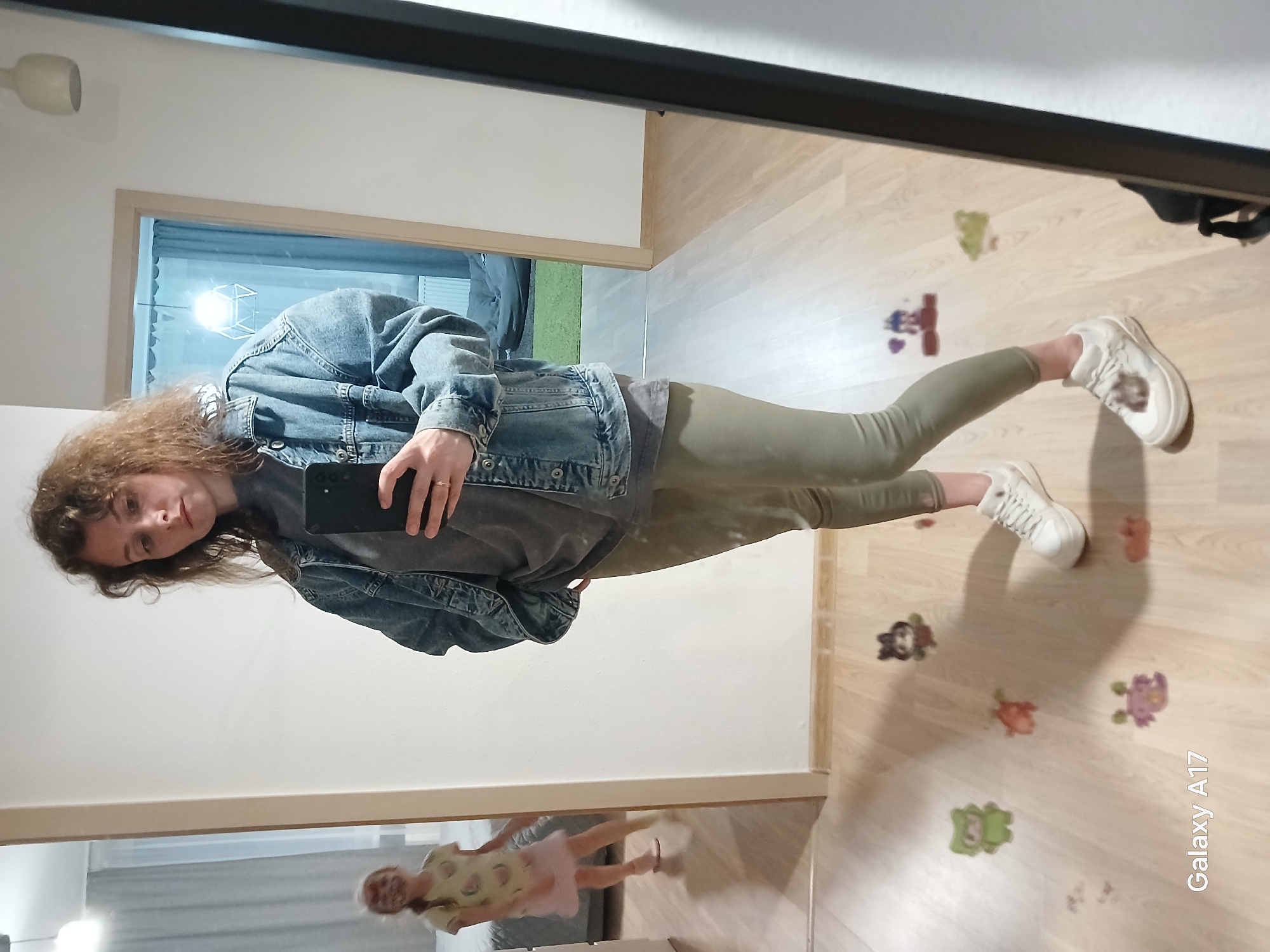 A mirror selfie inside a dwelling, likely a house or apartment. The person in the reflection is a Caucasian woman with curly hair, wearing a denim jacket over a dark-colored top and olive-green pants. She is holding a smartphone, likely taking the picture. The flooring is light-colored laminate with cartoon stickers scattered across it. A reflection of a child is seen near the bottom edge of the mirror. The environment includes a partially visible doorway and light-colored walls. The location is implied to be in Yekaterinburg, Russia.