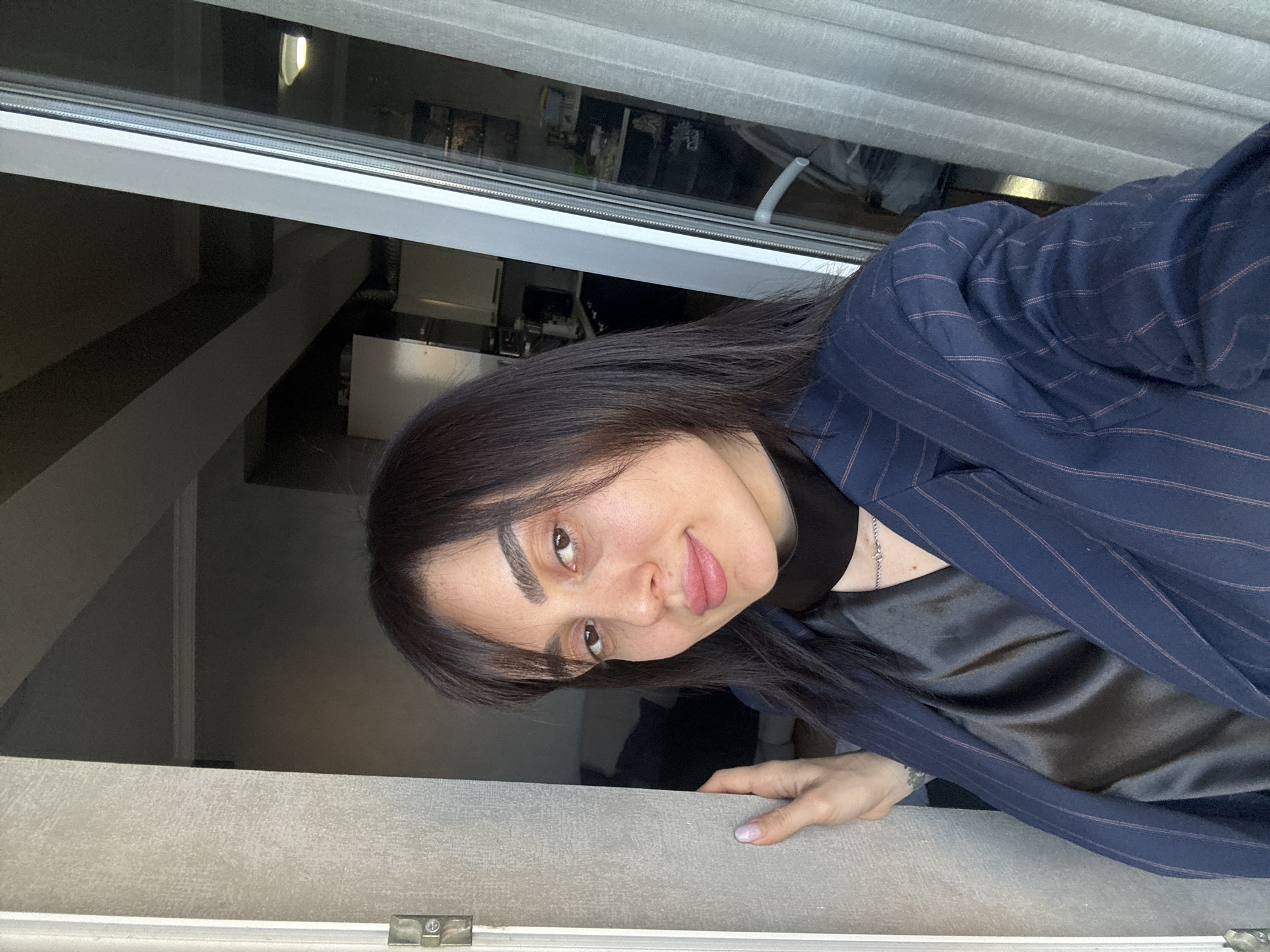 A woman with dark hair and fair skin is positioned in front of an open window. She is wearing a dark blazer with light pinstripes over a dark satin shirt. Her left hand rests on the window frame. Outside the window, glimpses of an urban scene can be seen. Based on visual architectural styles and the potential presence of former Soviet-era structures visible through the window, the location is likely in Tbilisi, Georgia.