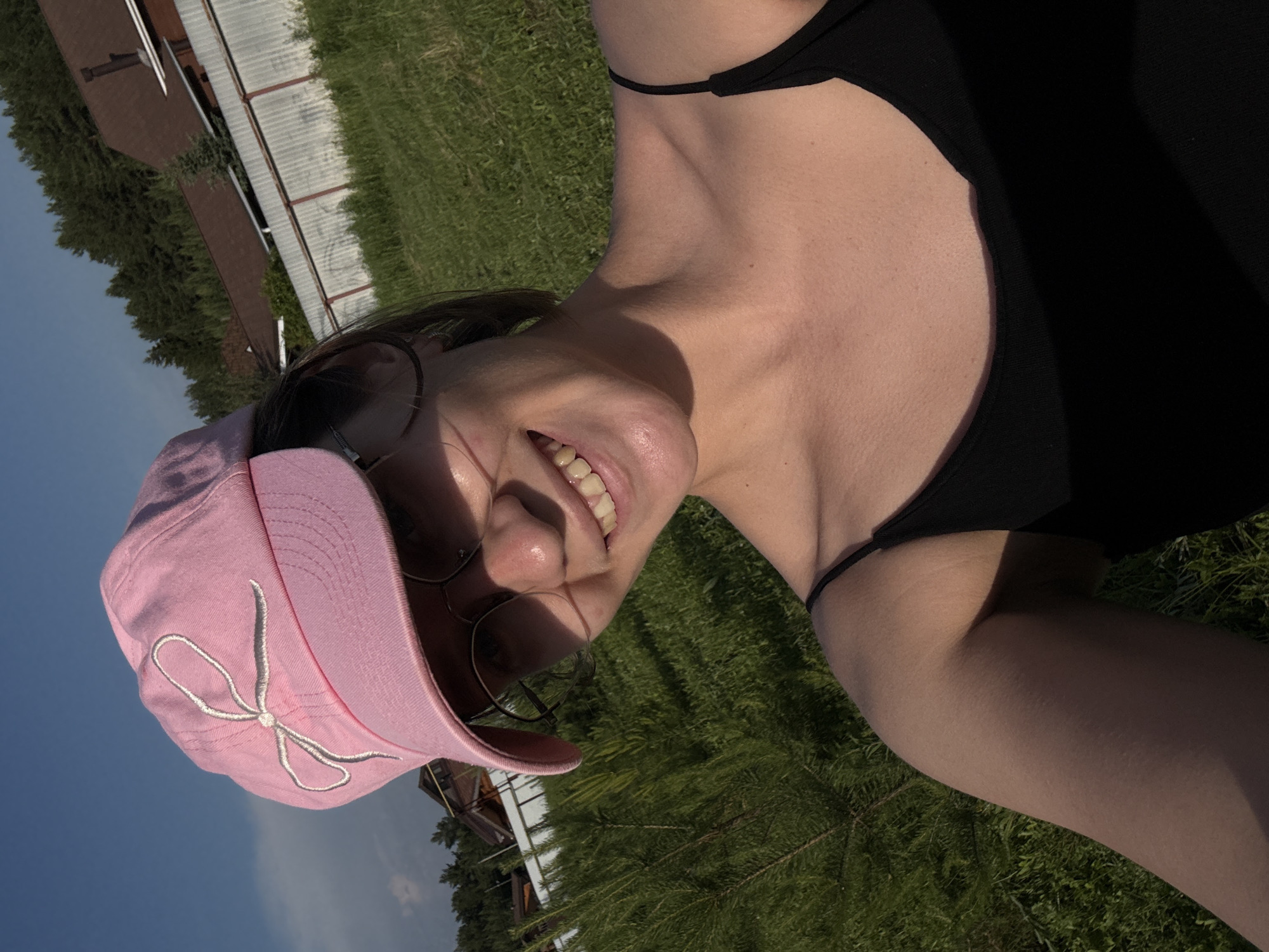 A selfie is taken from a low angle outside on a sunny day, with a young woman wearing a pink baseball cap with a white bow embroidered on it and glasses. She has a black tank top on and is smiling. In the background, there is a white metal fence, some trees, and houses with brown roofs. The ground is covered in green grass and some small pine trees. The sky is blue with a few clouds. The location is identified as Doksha, Russia.