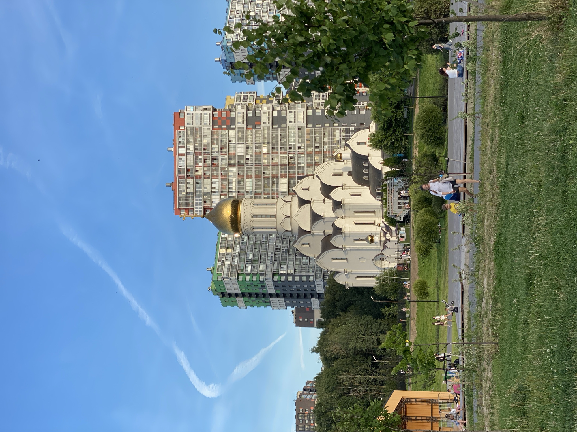 In the city of Krasnoborskoye, Russia, a white Orthodox church with a golden dome stands prominently against a bright blue sky with contrails. Surrounding the church are several tall, modern apartment buildings with colorful facades in shades of red, green, and blue. The foreground features a grassy park area with several people enjoying the outdoors. A woman and a young boy are walking, while others are sitting on benches or picnicking on the grass. A wooden structure, possibly a small stage or pavilion, is visible on the left side of the park. Trees and greenery are scattered throughout the scene, creating a peaceful urban park environment.