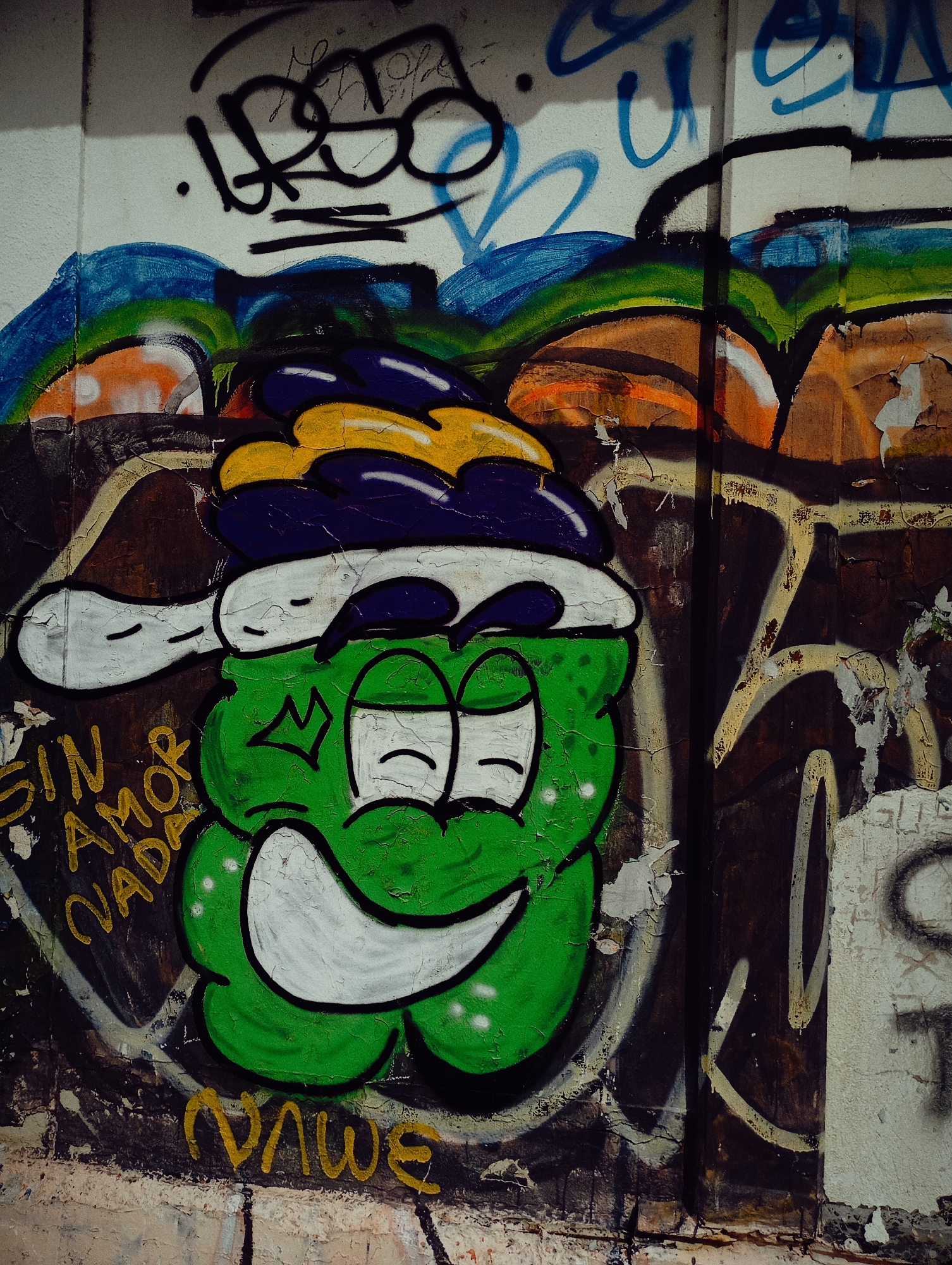 A section of a wall in Buenos Aires, Argentina, displays various layers of graffiti. A prominent cartoon-like figure, predominantly green with a white outline, features a relaxed expression with closed eyes and a broad smile. Above the figure, the graffiti includes colorful sections of blue, green, orange, yellow, and purple. A faded phrase "SIN AMOR NADA" is painted in yellow on the left. The letters "VAWE" are scrawled near the bottom. The wall surface shows signs of age with peeling paint and cracks. Other indiscernible graffiti surrounds the primary design. The overall atmosphere is urban and informal.